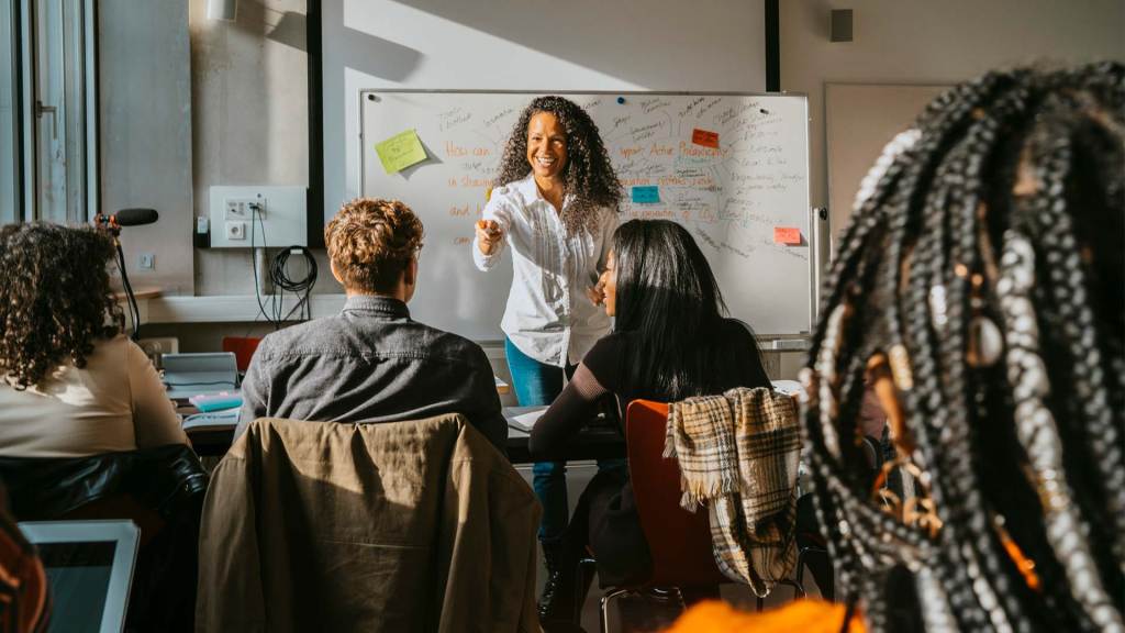 A professor giving a morning lecture to a diverse group of students. The professor is a woman with light brown skin and curly brown hair. She is smiling to the class.