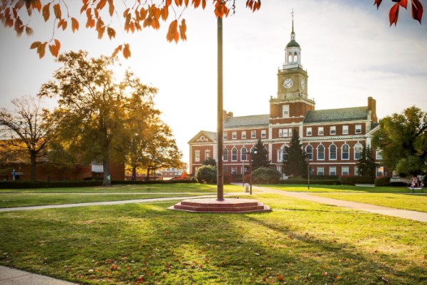 A historic brick building with a clock tower sits on a grassy, tree-lined campus under a bright sky. Autumn leaves frame the top of the image and sunlight filters through the trees.