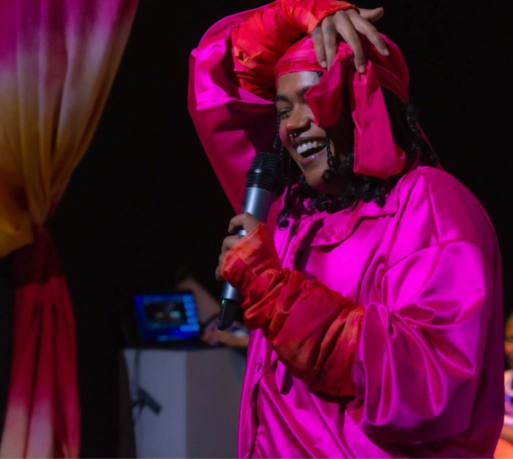 Smiling figure in fuschia satin shirt and head wrap, holding a microphone.