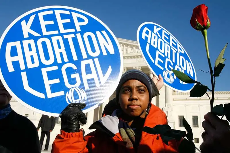 Local pro-choice activist Lisa King holds a sign in front of the US Supreme Court as a pro-life activist holds a rose nearby during the annual "March for Life". Lisa King is holding a royal blue sign with “Keep Abortion Legal” written in bold white font. She is wearing an orange jacket with a blue cap. 