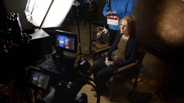 Gloria Steinem sitting on a film set in front of a camera with a bright light shining on her. 