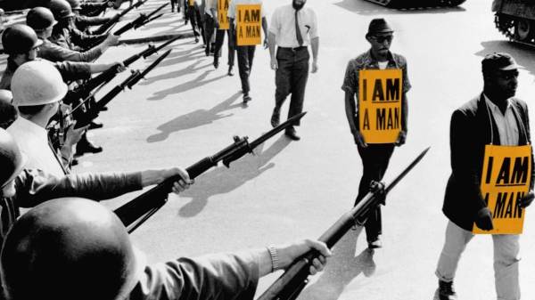 A black and white photo of Black men with signs that read "I AM A MAN" marching in front of armed military personnel 