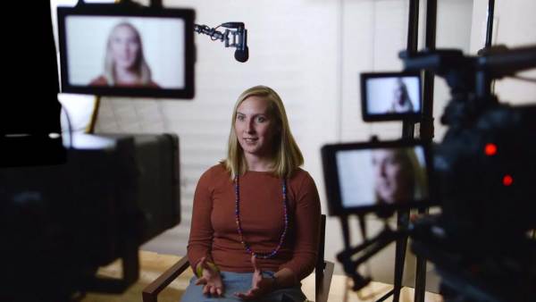 May Boeve sitting on a film set in front of multiple cameras with her face displayed on all monitors. 