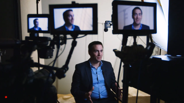 Michael Clemens sitting on a film set in front of multiple cameras with his face displayed on all monitors. 