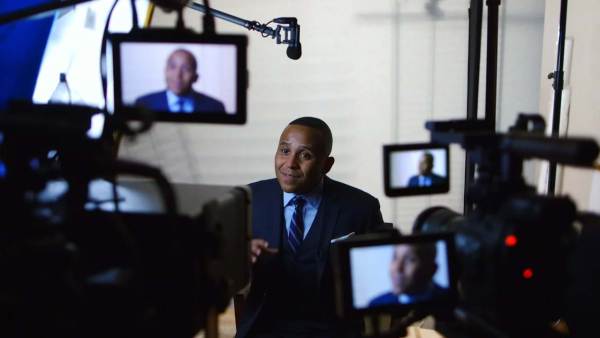 Rashad Robinson sitting on a film set in front of multiple cameras with his face displayed on all monitors. 