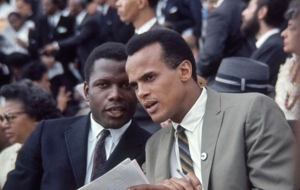 Sidney Poitier, in a navy suit, leans in to speak to Harry Belafonte, in a gray suit, in a crowd. Belafonte is holding a cigarette in one hand and a stack of papers in the other. 