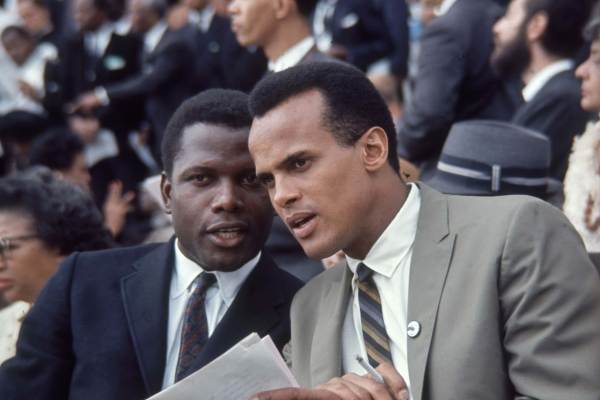 Sidney Poitier, in a navy suit, leans in to speak to Harry Belafonte, in a gray suit, in a crowd. Belafonte is holding a cigarette in one hand and a stack of papers in the other. 