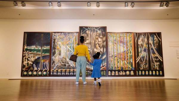 A woman and child stand before a five-paneled painting in a gallery. The panels each depict abstract scenes of bridges in fantastical colors.