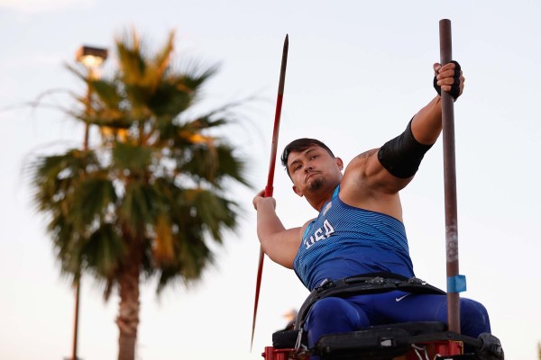 As late day light illuminates palm trees, Paralympian Justin Phongsavanh, leaning back on a wheelchair, prepares to throw a javelin during a competition in Arizona.