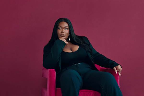 Before a vibrant magenta backdrop, Jordyn Jay, a Black trans femme, leans elegantly on a plush, pink chair, her thoughtful pose accented by a hand delicately supporting her chin. Draped in a sleek, black pants suit, her long hair cascades from a central part, providing contrast to the surrounding hues.