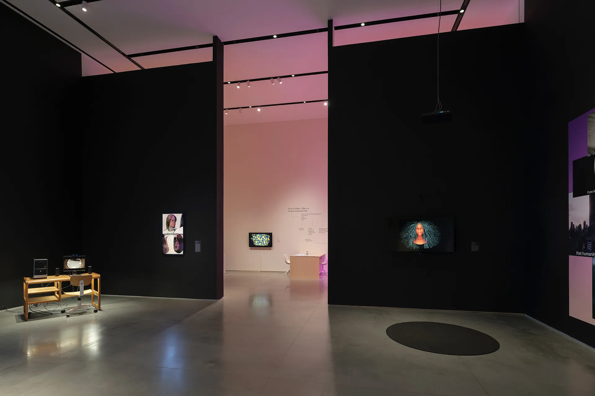 Two black walls with an aisle down the middle. Each wall has a video monitor in the center. Standing on the floor in front of the left wall, there is a wooden desk with a computer on it. On the floor, in front of the right wall, there is a round black rug.