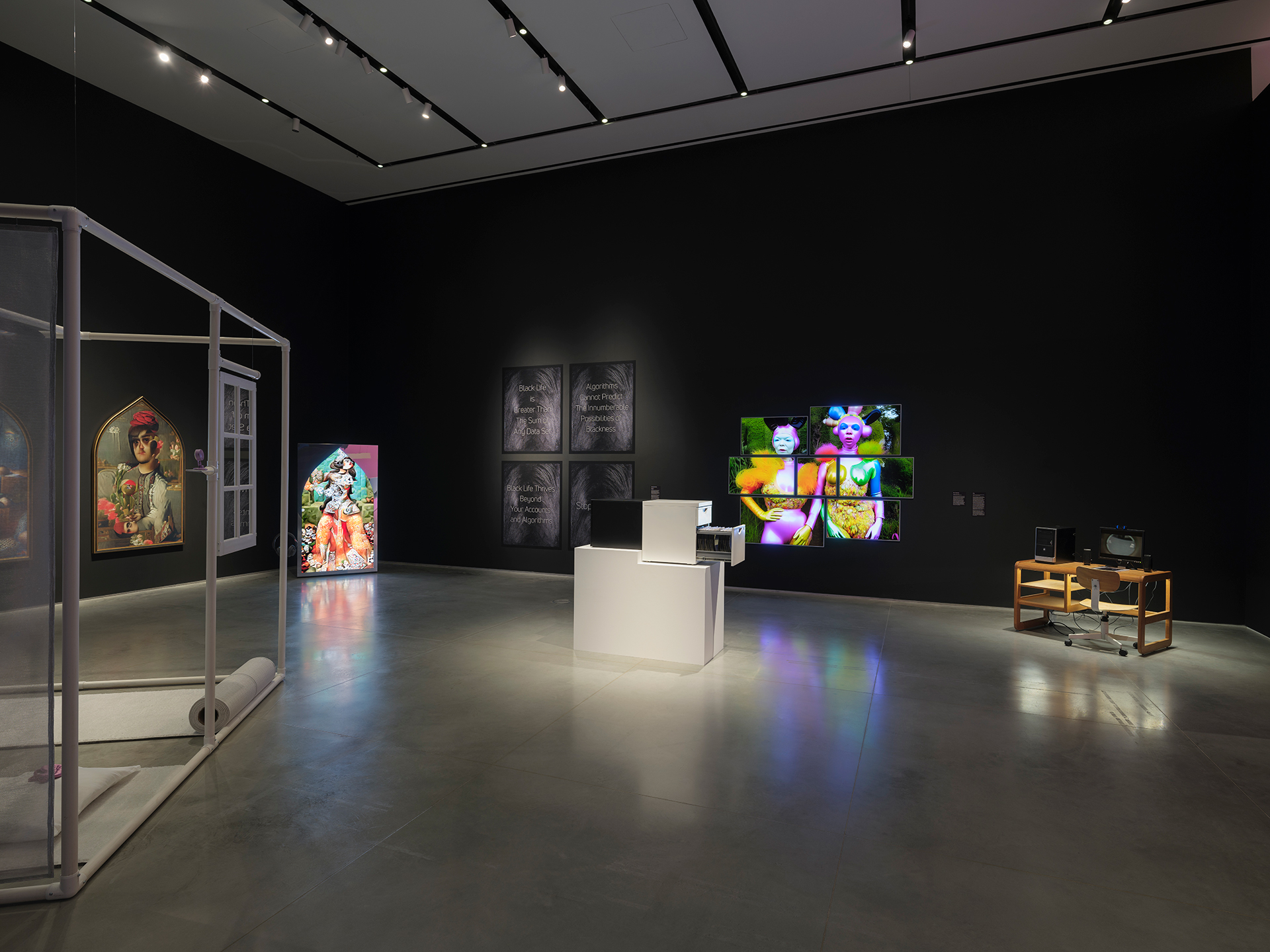 Gallery interior with black walls and a gray floor. There are several artworks displayed including freestanding works on a desk and one on a pedestal, as well as mounted works on the walls, some of which are displayed on screens.