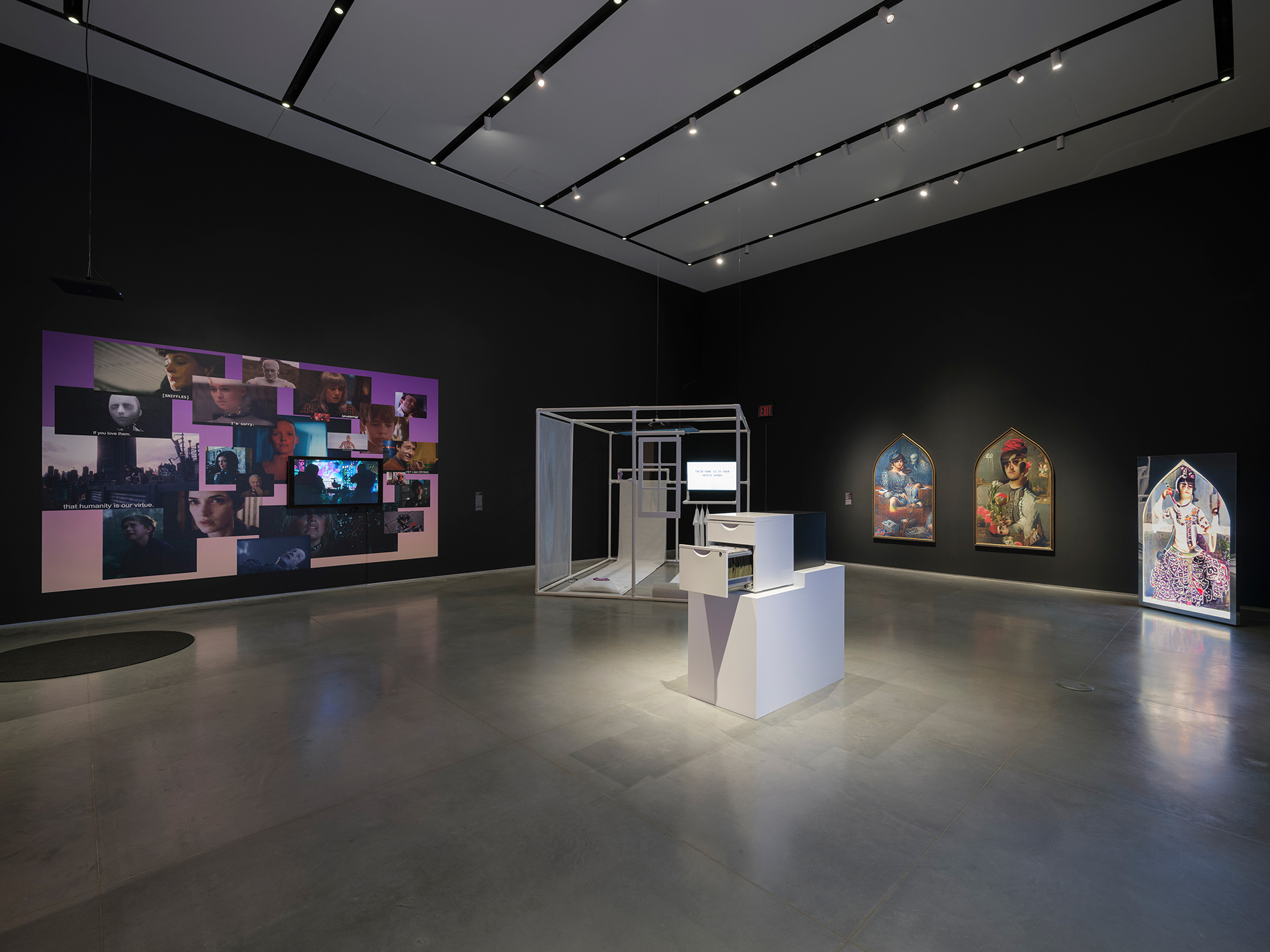 Interior view of a gallery space where two black walls meet at a corner. There are mounted artworks on both walls. The floor is gray and features two additional freestanding artworks, one is on a pedestal and the other is a large cube made of PVC pipes.