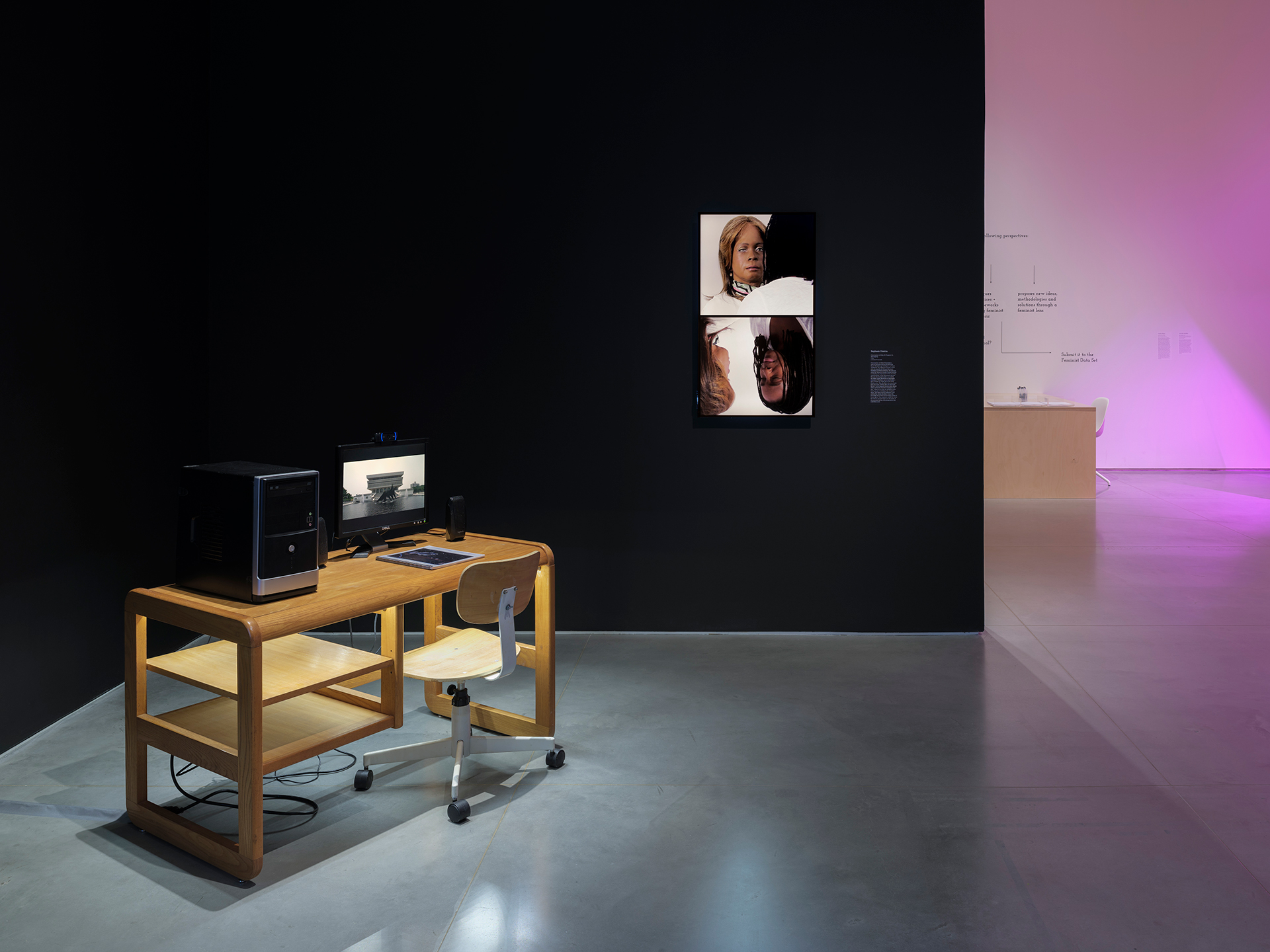 Gallery interior with a black wall and a gray floor. To the left, a wooden desk with a matching wooden chair holds a computer, a monitor, and a packet of paper. Mounted on the wall, to the right of the desk, a screen displays images of two women.