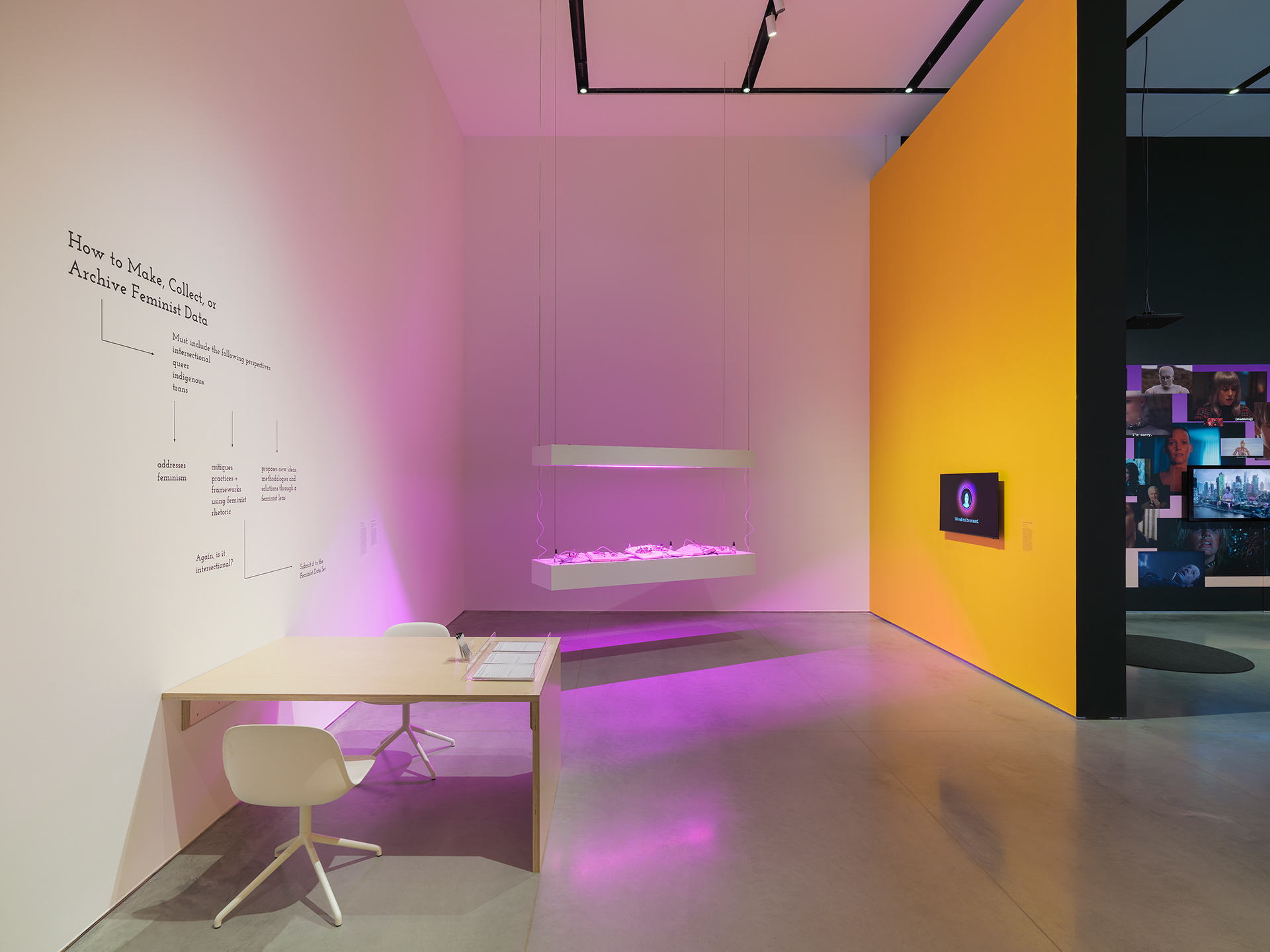 Interior space with three walls. The left and back wall are white, the right wall is yellow with black trim. There is a wooden desk with white chairs pushed against the left wall and two hanging platforms hovering near the back wall are glowing purple.