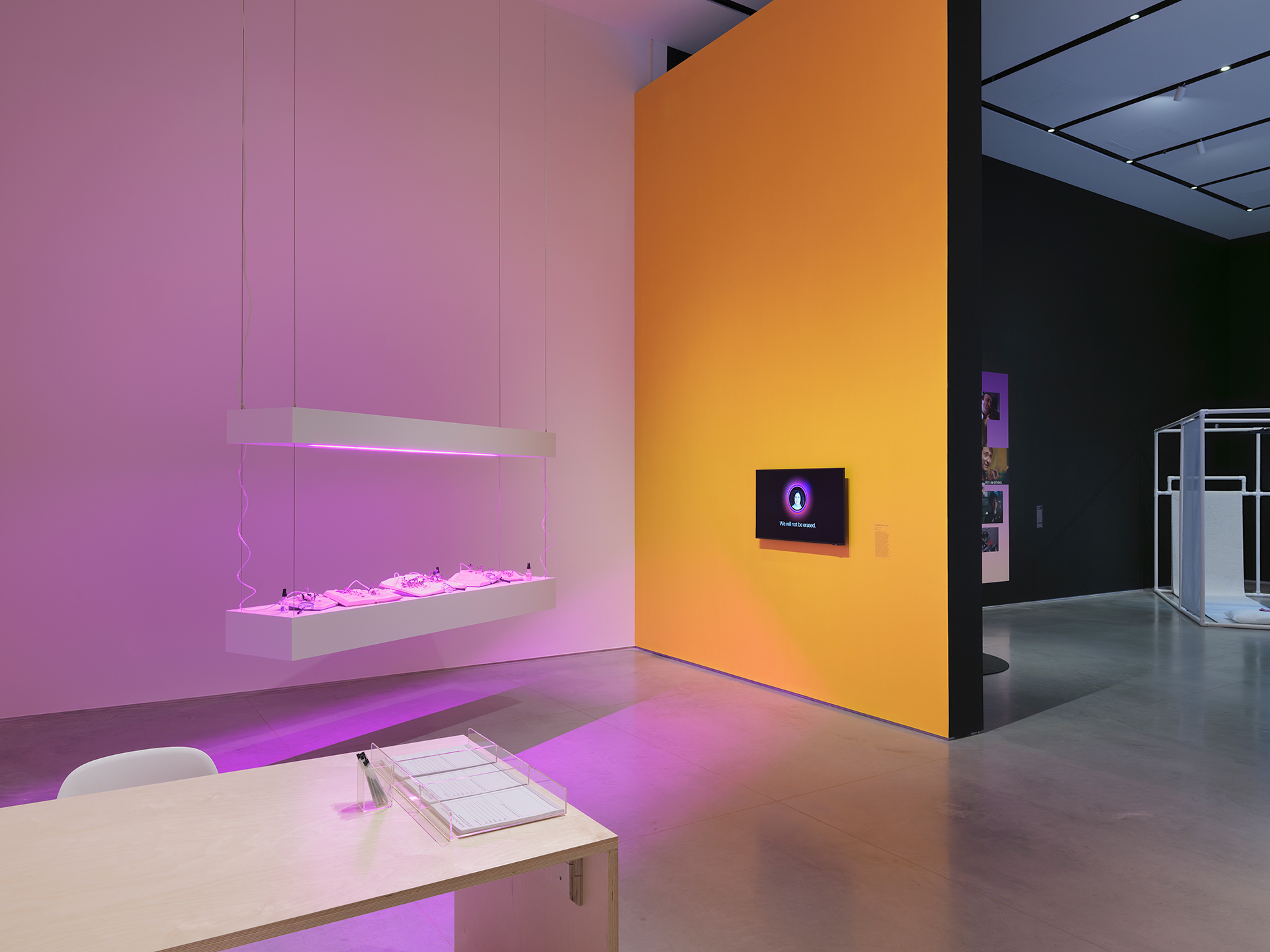 Gallery interior with two walls bisected by a partial wall. The wall on the left is white and the wall on the right is black. The bisecting wall is yellow with black trim and holds a video monitor. Hanging from the ceiling in front of the white wall are two narrow platforms hovering parallel to one another. There is a purple glow on the left side of the room.