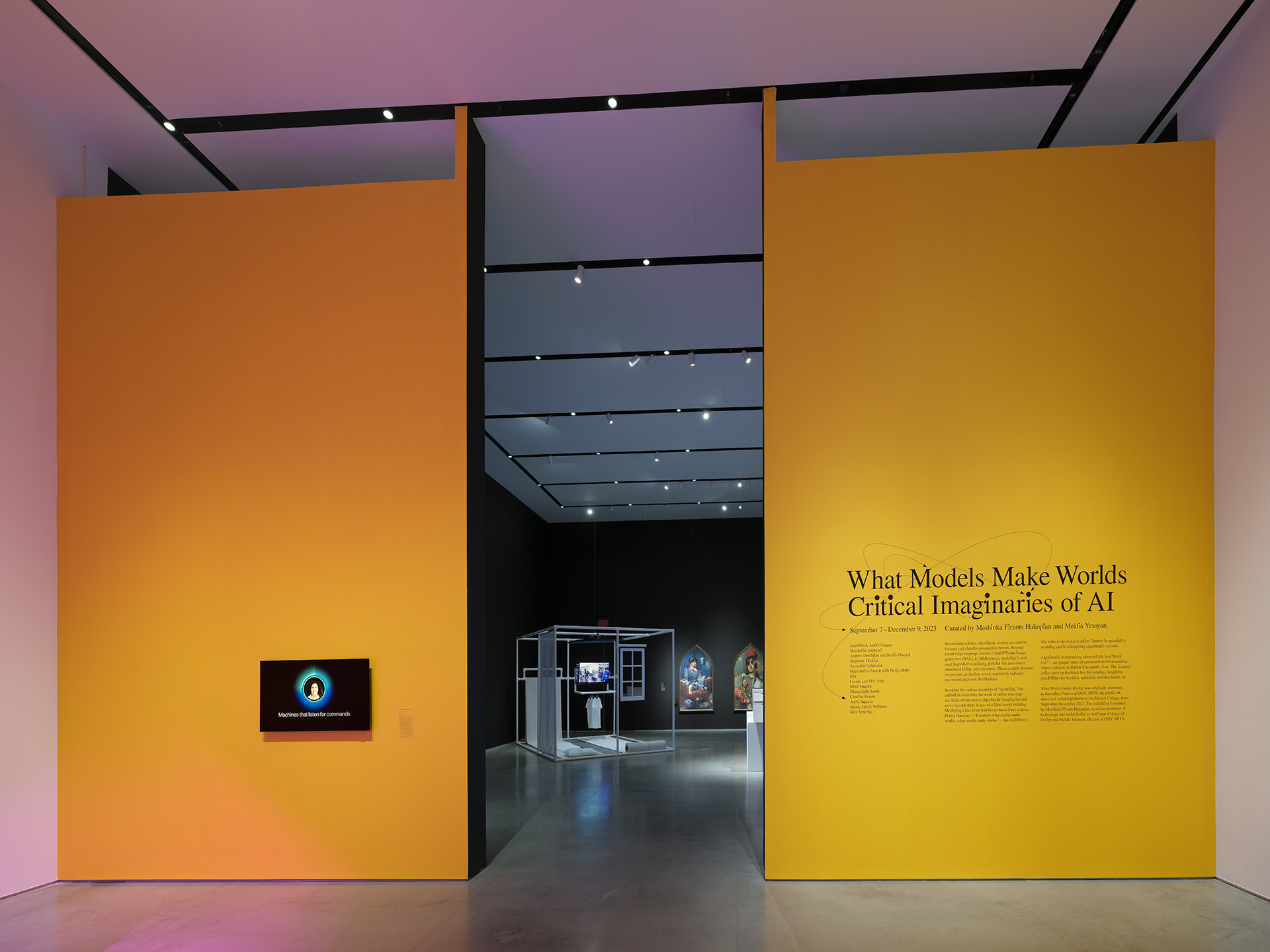 Interior of an art gallery featuring two yellow walls with an aisle in between. The entry text on the right wall reads “What Models Make Worlds, Critical Imaginaries of AI.” Further down the aisle between the walls, some artworks can be seen deeper inside the gallery.