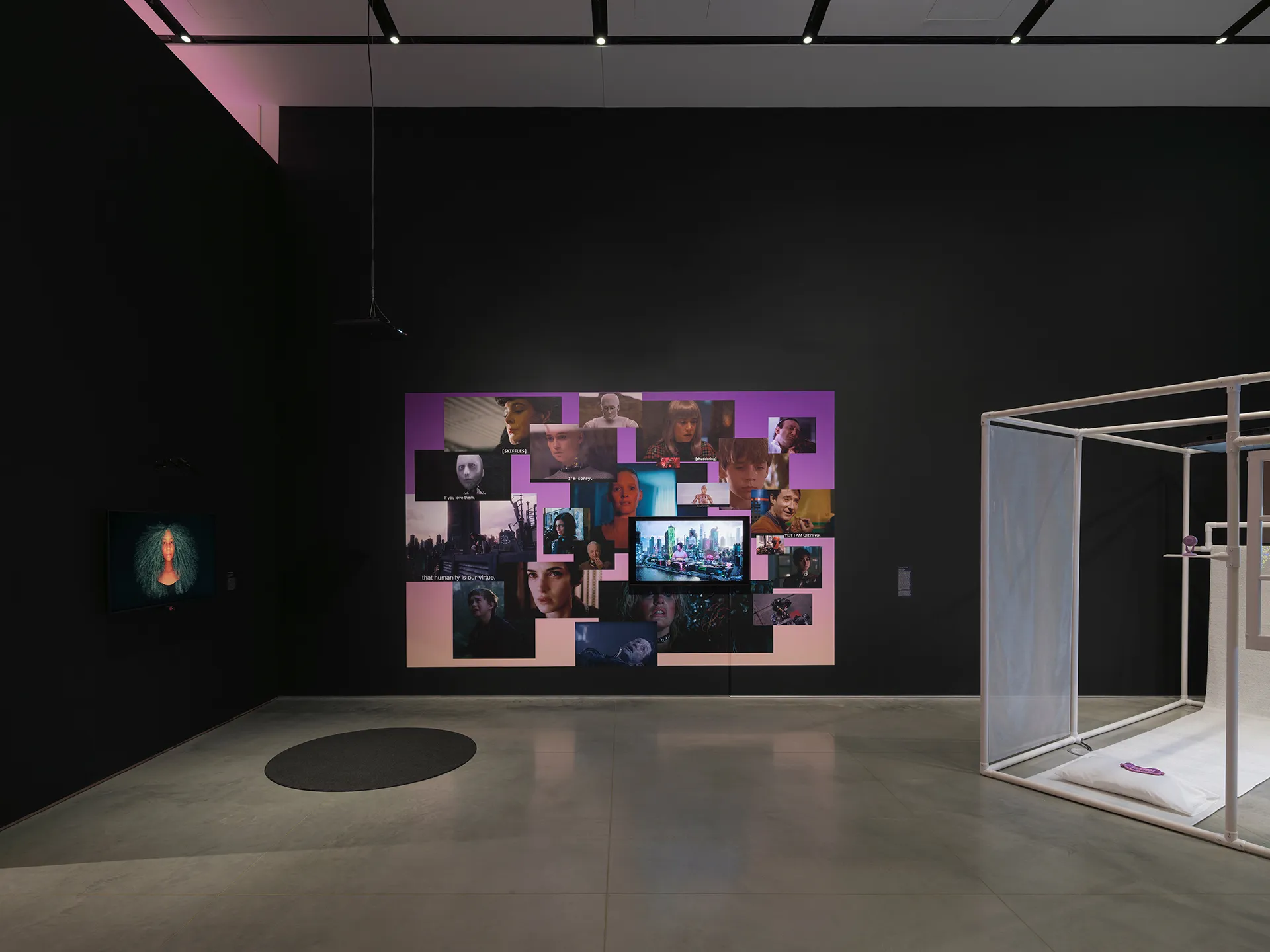Gallery interior with black walls and a gray floor. On the back wall, there is a print collaged with images from popular media. On the right, there is a partial view of a structure made of PVC pipes. On the left, at an angle, a mounted video monitor displays a woman’s face. There is a round rug on the floor.