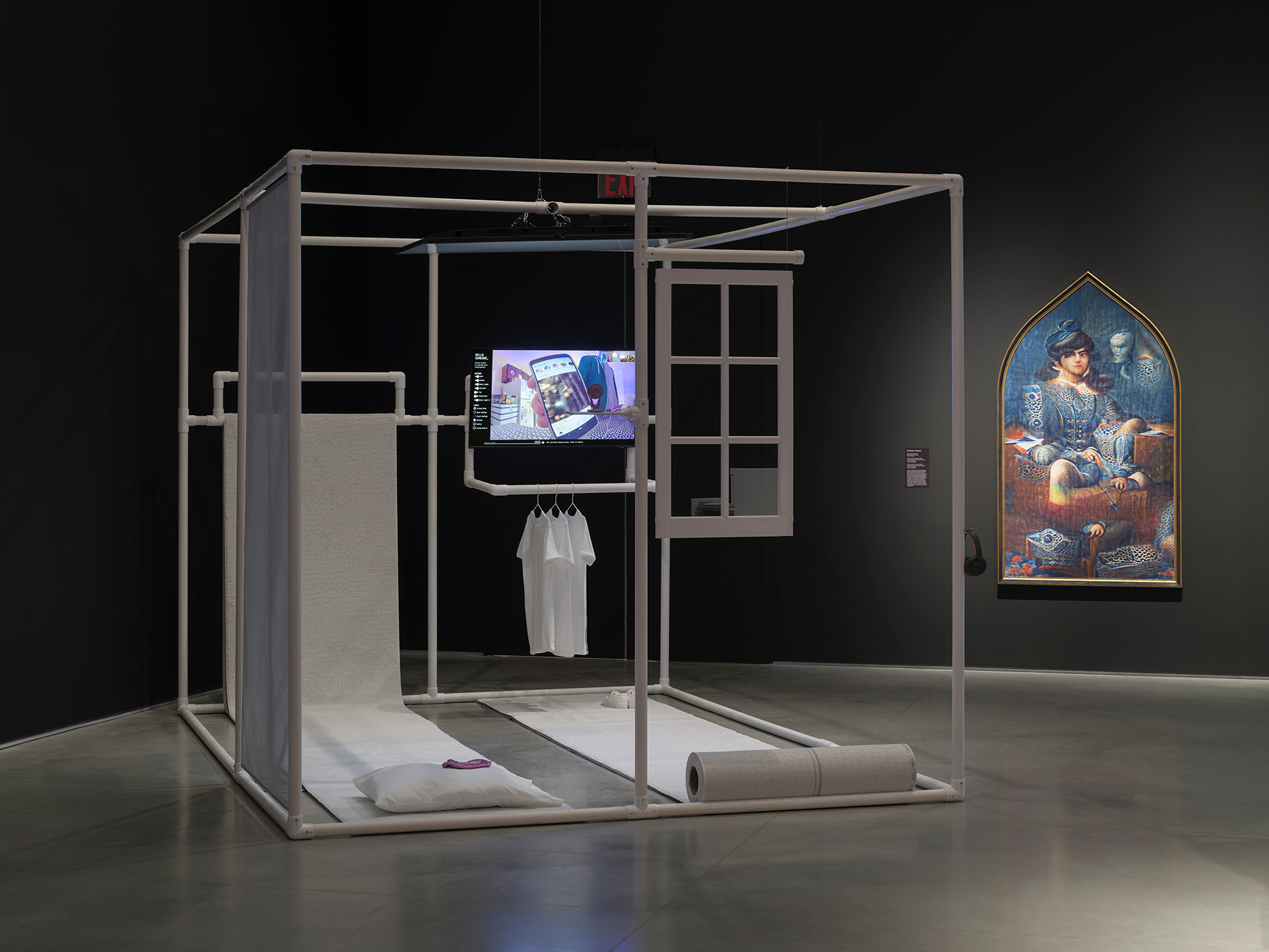 Gallery interior with a black wall and a gray floor. There is a large cube made of PVC pipes in the center of the floor. It has two rugs running across the floor, contained inside the borders of the PVC pipe frame. One horizontal pipe holds white t-shirts on hangers. There is a television monitor embedded inside the PVC pipe frame. On the wall to the right of the cube, there is a framed portrait.