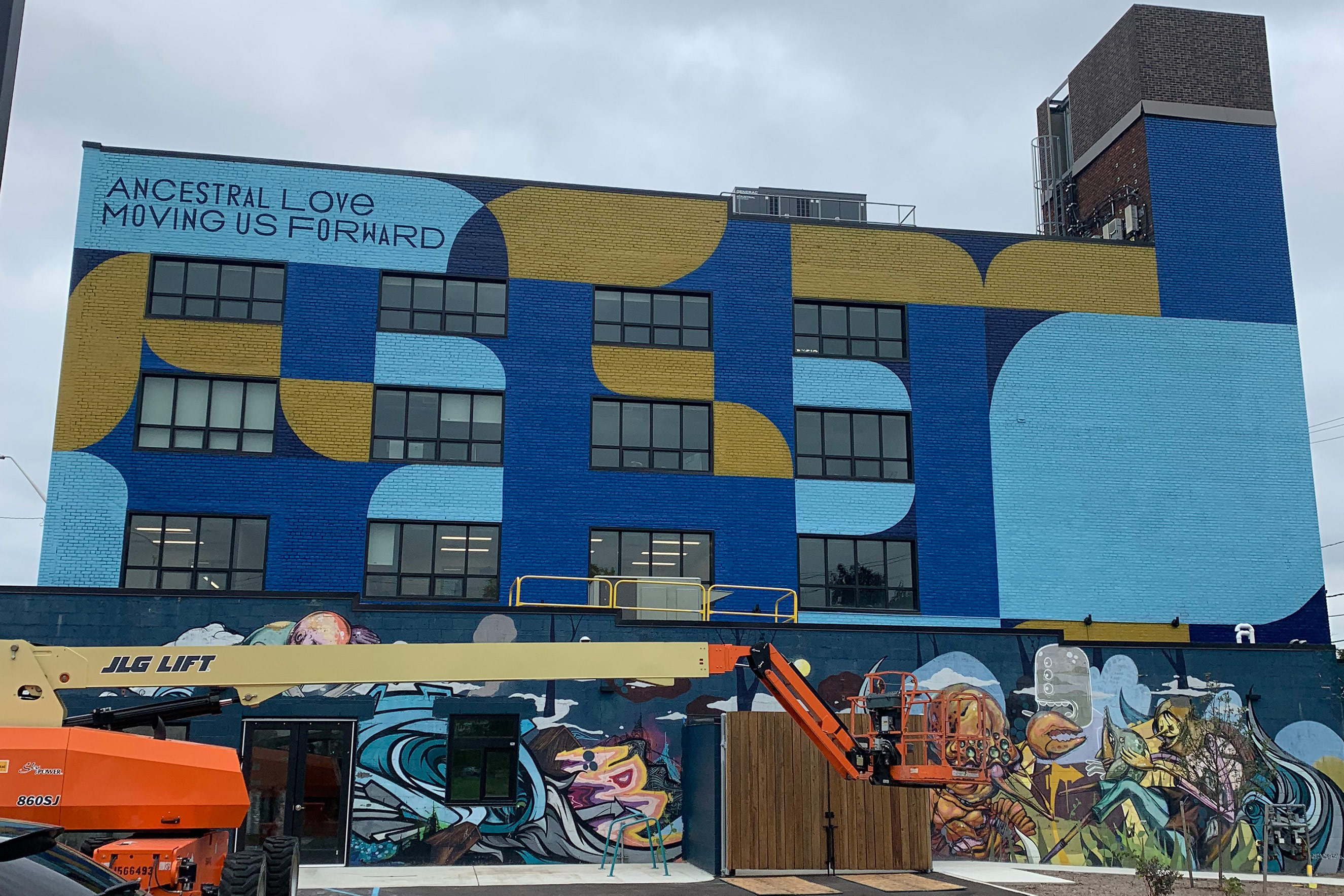 Mural in Detroit, a city in the United States, that features blue and gold geometric shapes and the text, "ANCESTRAL LOVE MOVING US FORWARD." A second detailed mural is on the ground level.