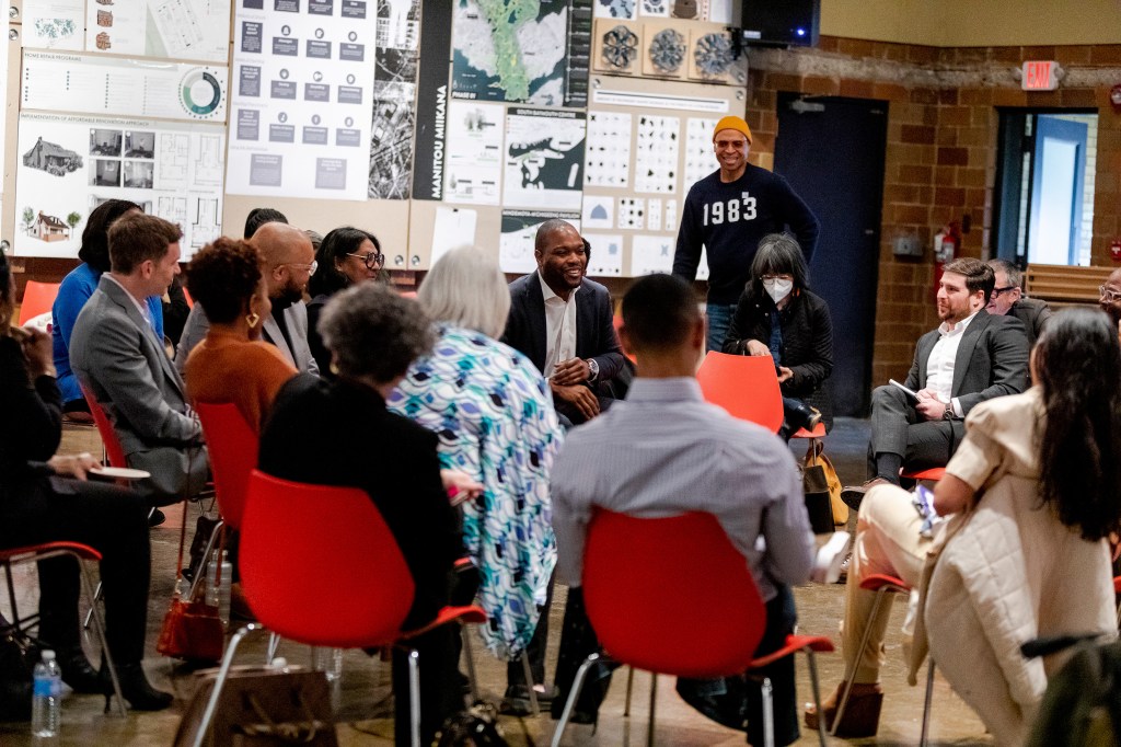 A group of 15 people sit in a cluster of red chairs in a brick walled room with maps and charts on the wall. They are being addressed by a dark skinned man in a yellow meaning cap and a black t-shirt labeled 1983.