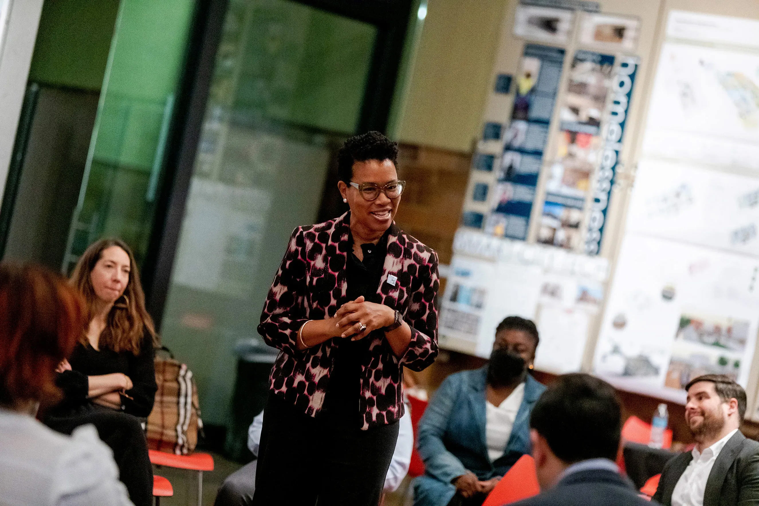 A smiling dark skinned woman wearing a patterned pink and black blazer speaking in the middle of a circle of people sitting in red chairs.
