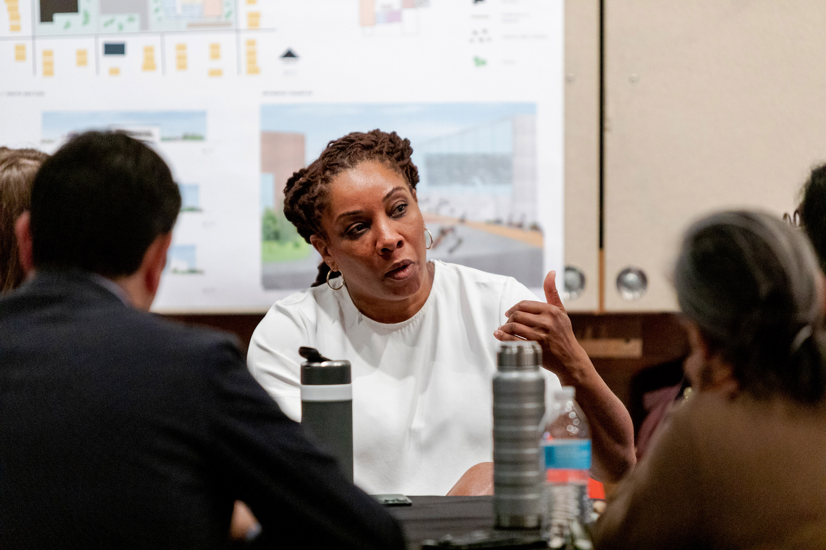 A dark skinned woman wearing a white t-shirt speaking at a conference table to others. Two water bottles are in front of her.