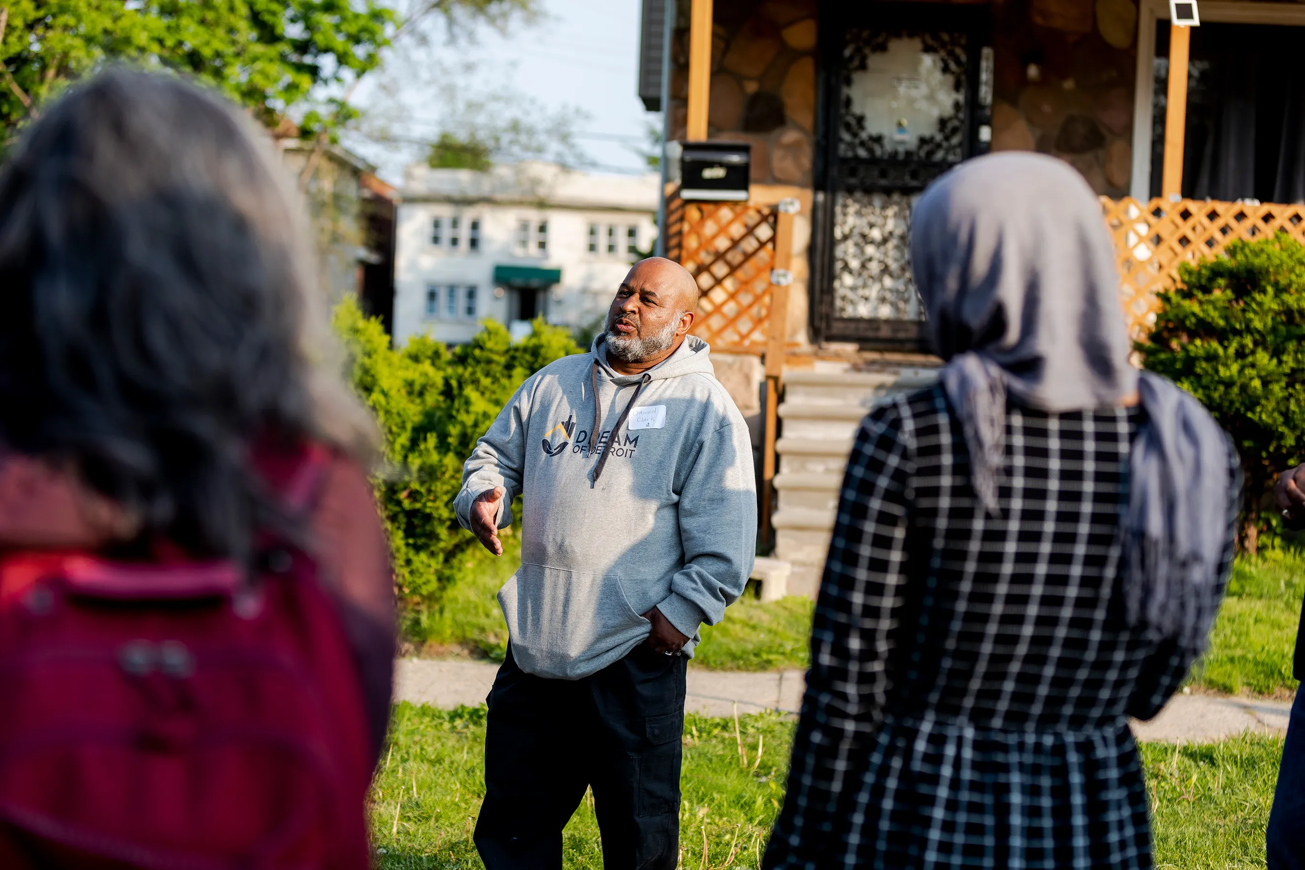 A dark skinned man wearing a gray sweatshirt is speaking to two people in front of a house with a brown porch.