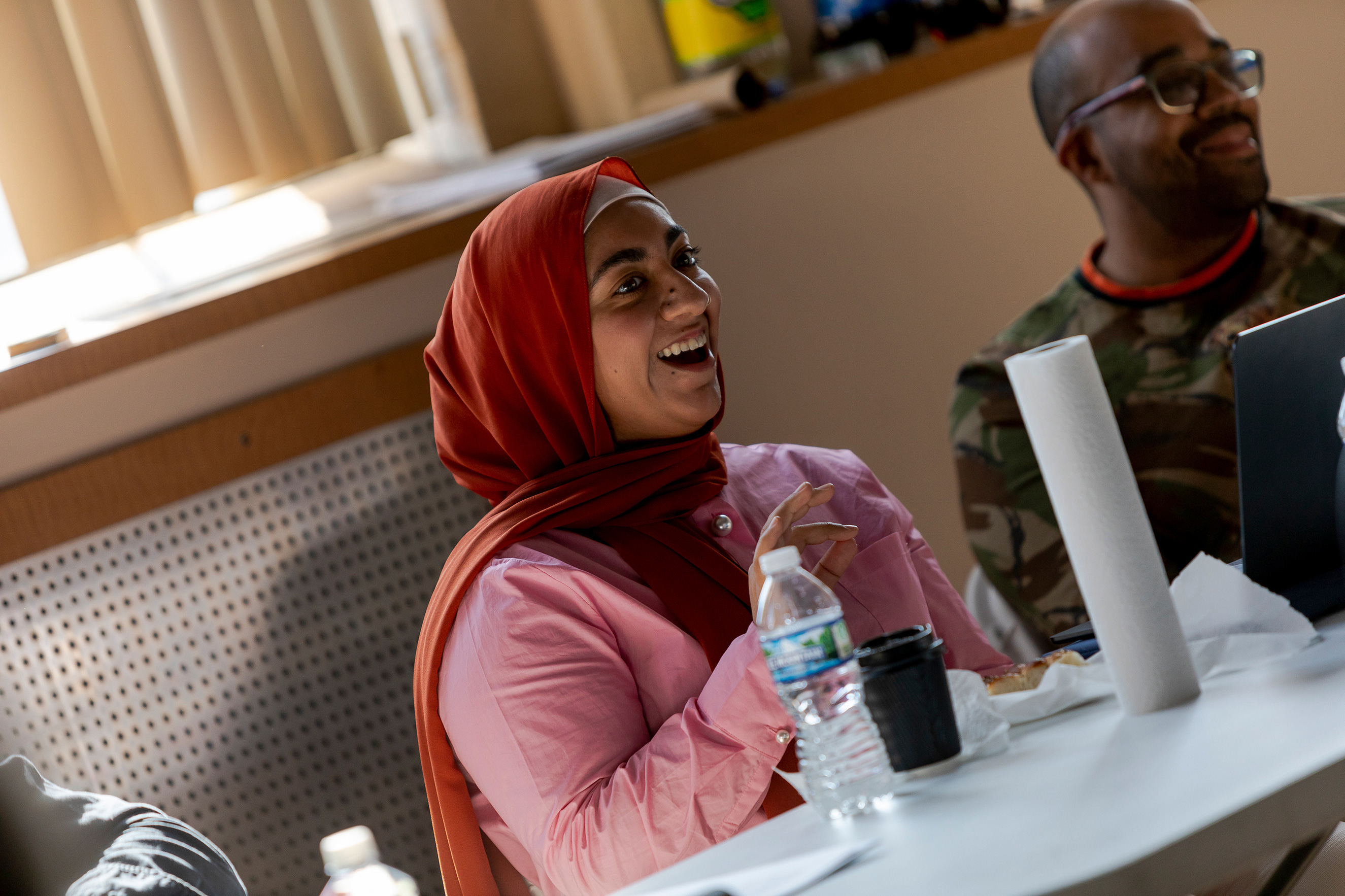 A light skinned woman wearing a pink blouse and a red hijab is speaking with joy.
