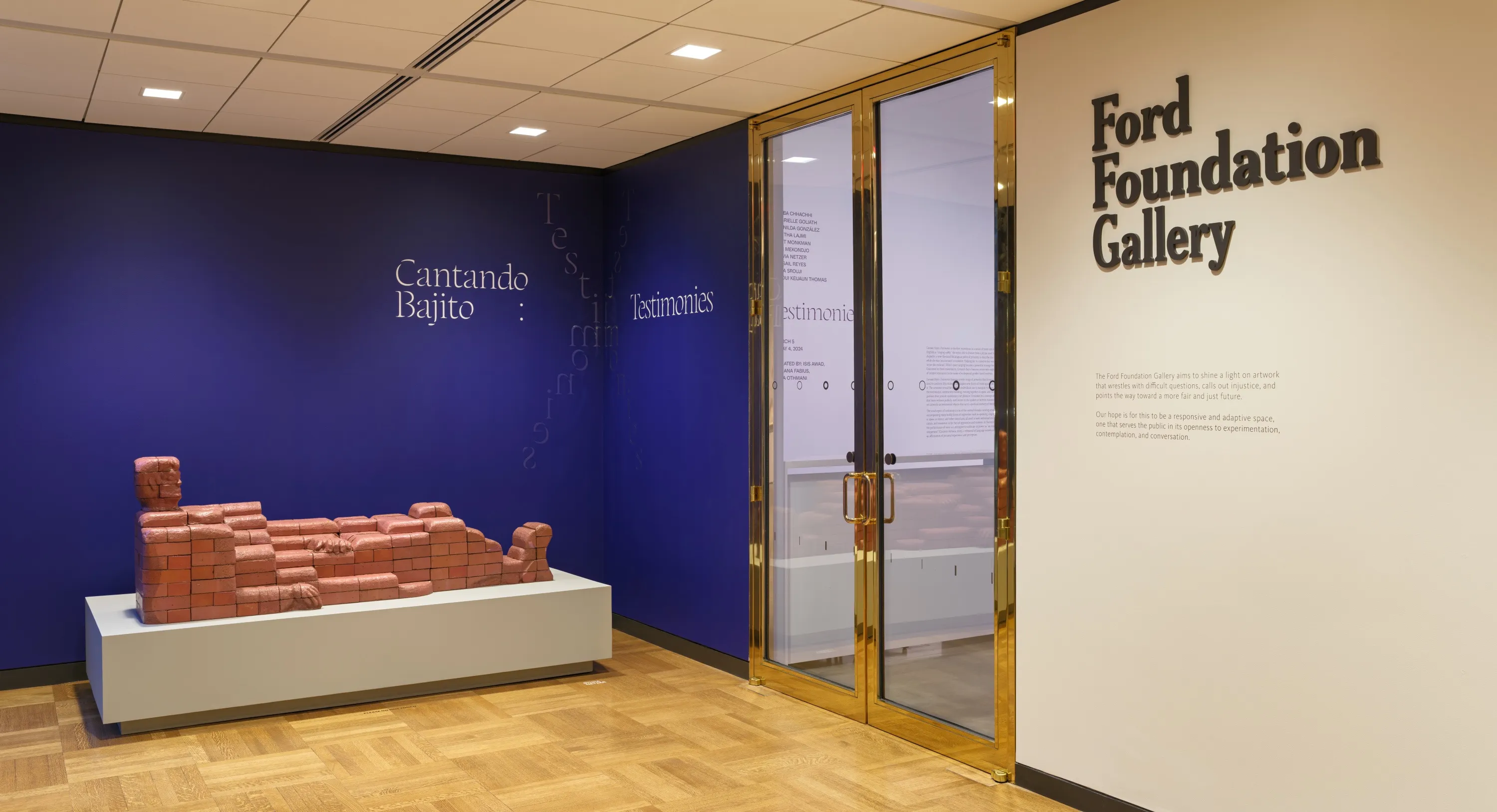 An installation of a brick-lady sculpture on a gray pedestal in front of a dark purple wall with text “Cantando Bajito: Testimonies”. To the right of the wall is a gold-metallic door frame; and a white wall with some text, titled with the header “Ford Foundation Gallery”.