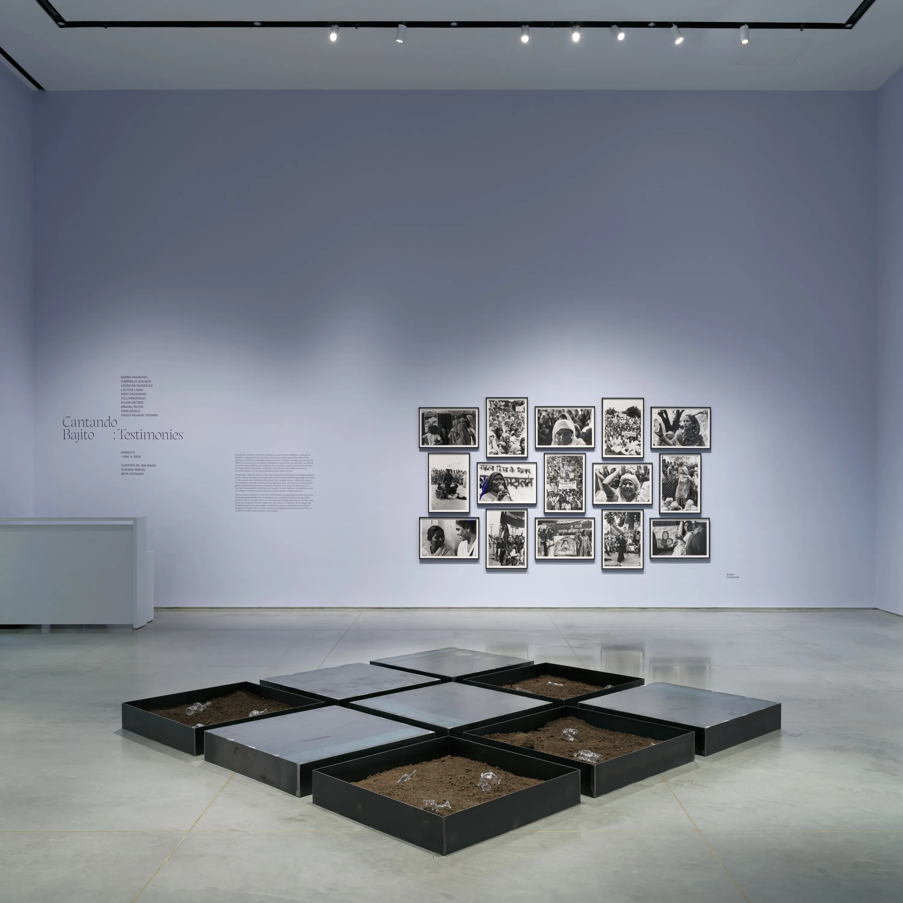 Gallery interior with light lavender walls and a gray floor. A three by three steel-tray installation sits in the middle of the floor. In the background above a white desk, the wall has text with header “Cantando Bajito: Testimonies”. To the right is an arrangement of fifteen framed black and white photographs.