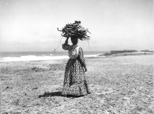 A black and white photograph of a woman standing in a field of dirt and a body of water in the far background. She is walking towards the opposite direction of the camera, in a long floral-like patterned dress with long sleeves. A piece of cloth is tied diagonally across her shoulder, and she is balancing what appears to be a bundle of branches on top of her head.