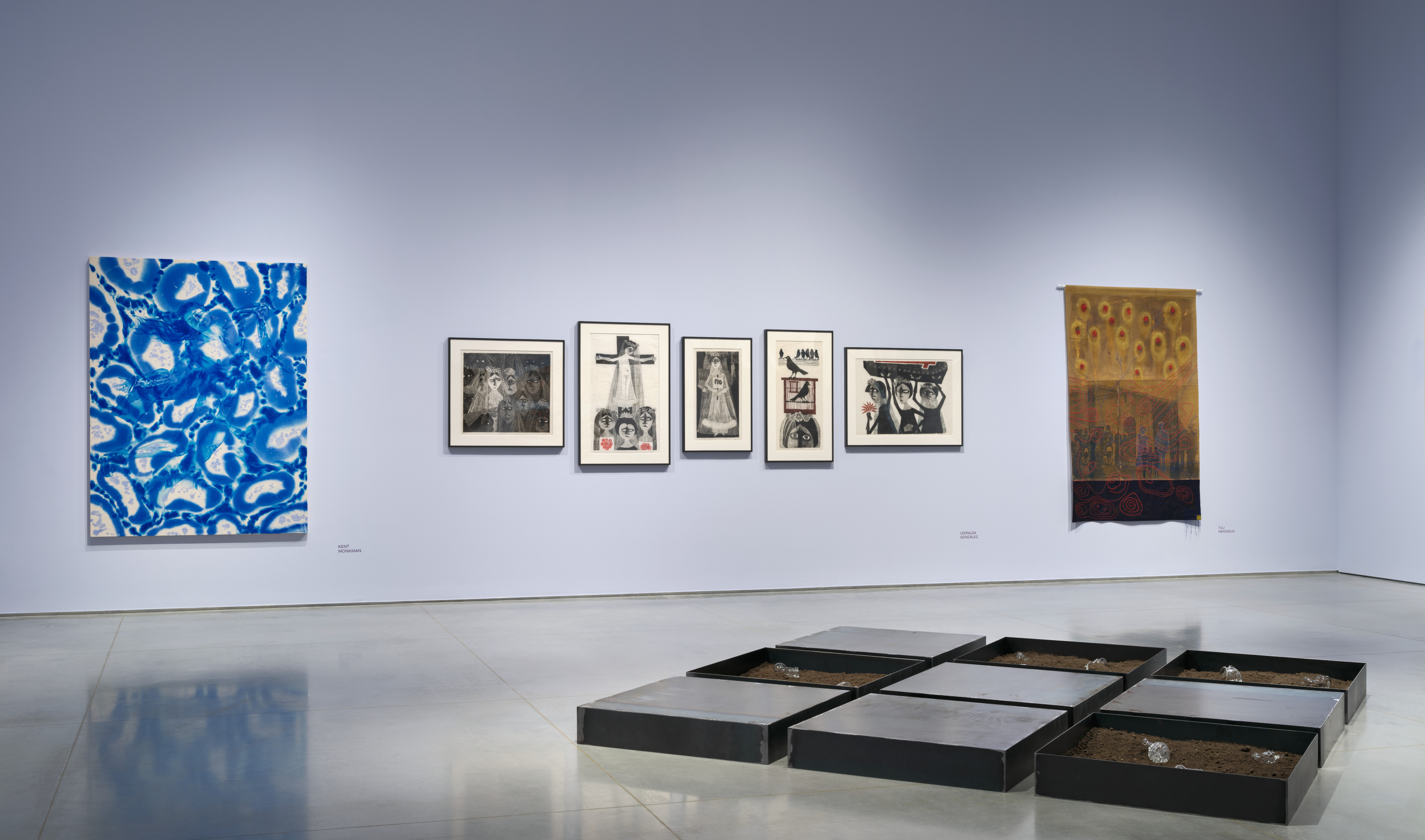 Gallery interior with light lavender walls and a gray floor. On the floor is a three by three steel-tray installation. On the wall are several artworks in a line. From left to right, one blue and white patterned painting, then five framed woodcuts, then a brown canvas with various painted and embroidered elements.