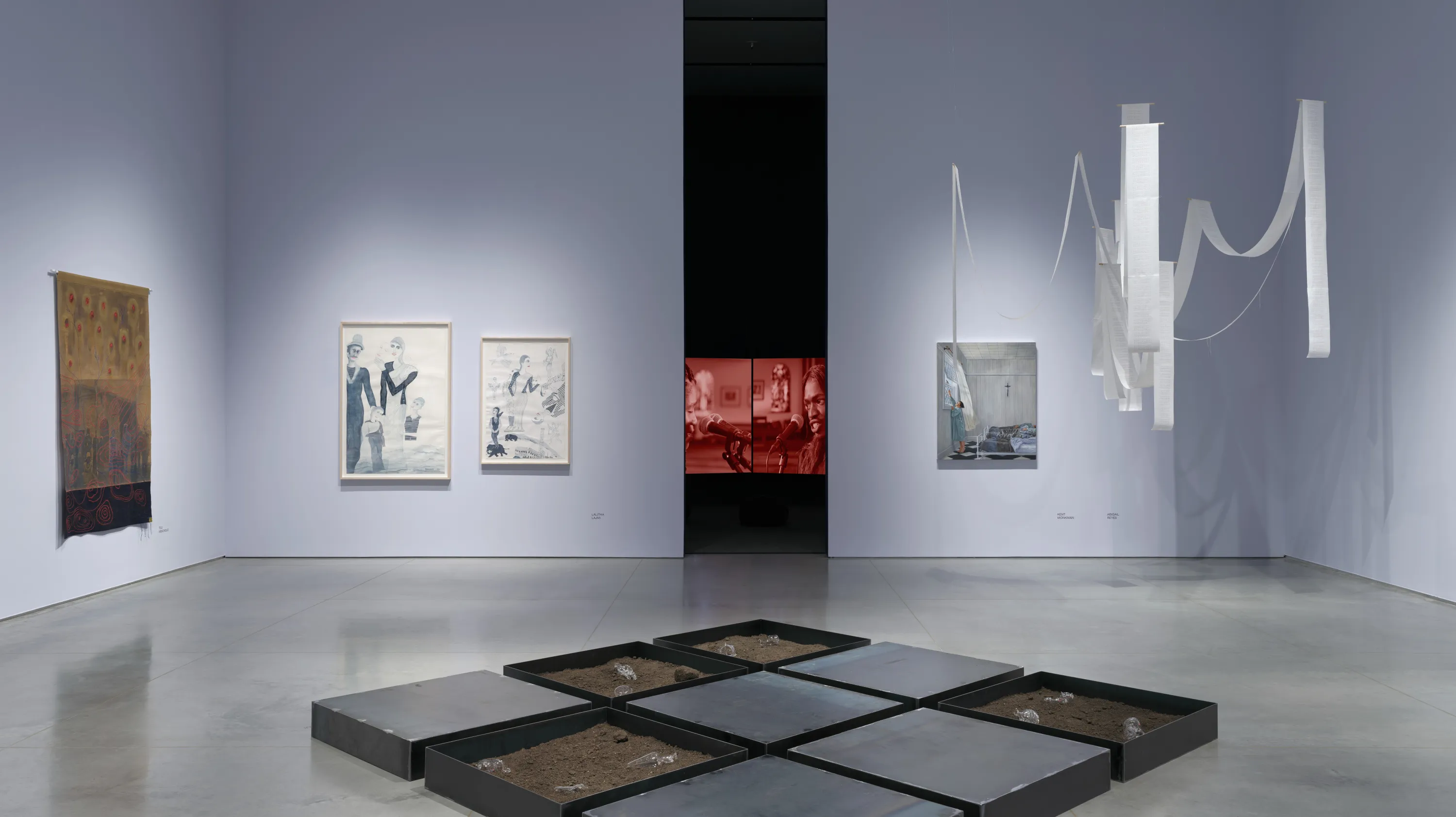 Gallery interior with two light lavender walls with an aisle down the middle leading to a dark room with a video installation. On the left wall is a brown canvas artwork, and two framed watercolor paintings. On the right wall is an acrylic painting, and an artwork installation of long ribbons of white paper hanging vertically and in dipped arcs. On the floor is a three by three steel-tray installation.