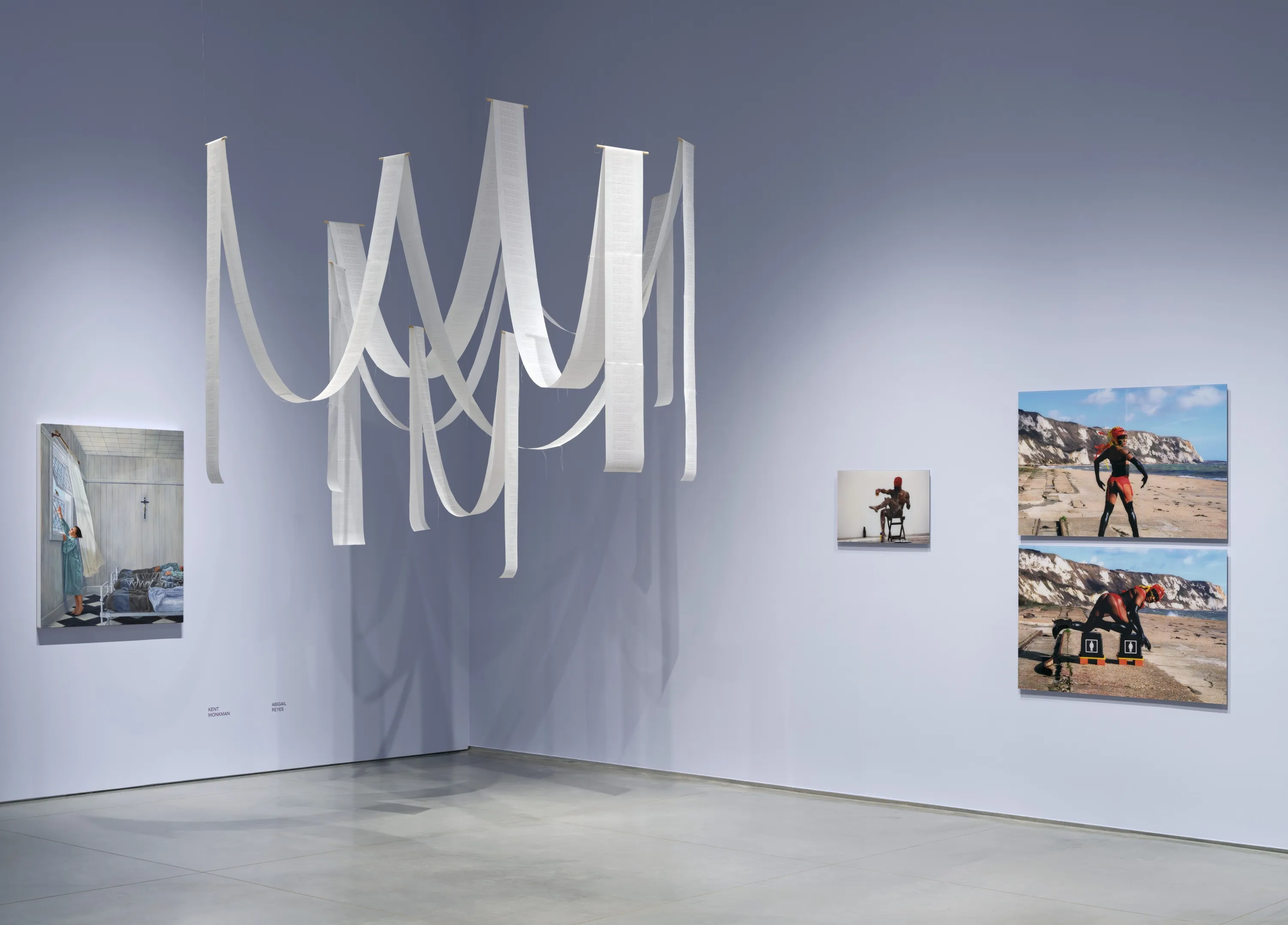 Gallery interior with light lavender walls and a gray floor. An acrylic painting is mounted on the left wall. And an artwork installation of long ribbons of white paper is hanging vertically and in dipped arcs in the middle. And on the right wall, are three photographs mounted on the wall - the photos have a central Black femme person in different poses.