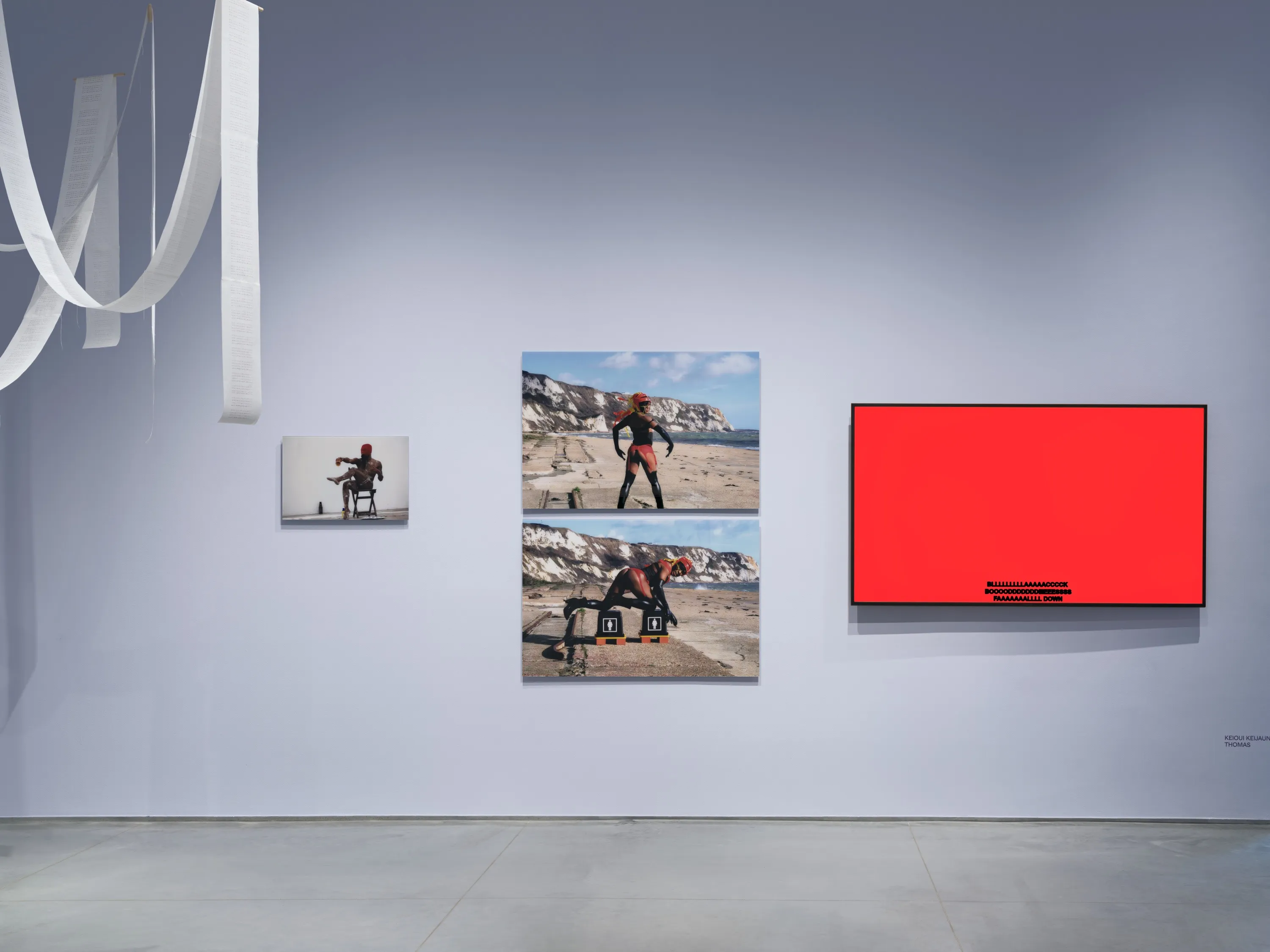 Gallery interior with light lavender walls and a gray floor. A part of an artwork installation of long ribbons of white paper is hanging to the left, and on the wall are three photographs mounted on the wall depicting a Black femme person in different poses. And to the right is a large monitor showing a bright red screen with black text captions at the bottom.