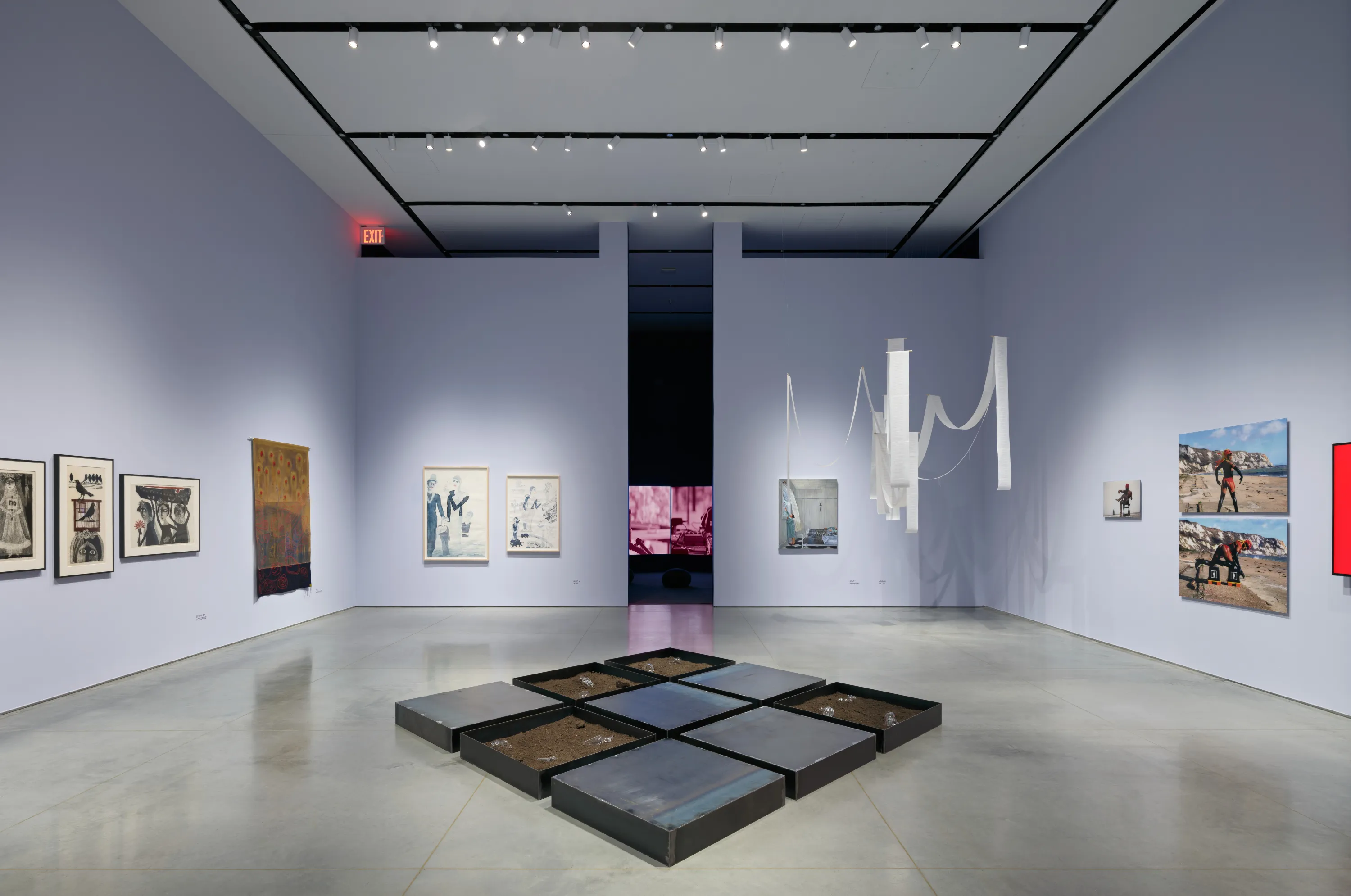 Gallery interior with a gray floor and light lavender walls with an aisle down the middle leading to a dark room with a video installation. There are several artworks displayed including paintings, photographs, canvas mounted on the wall and a hanging white paper ribbon installation and steel tray installation on the floor.