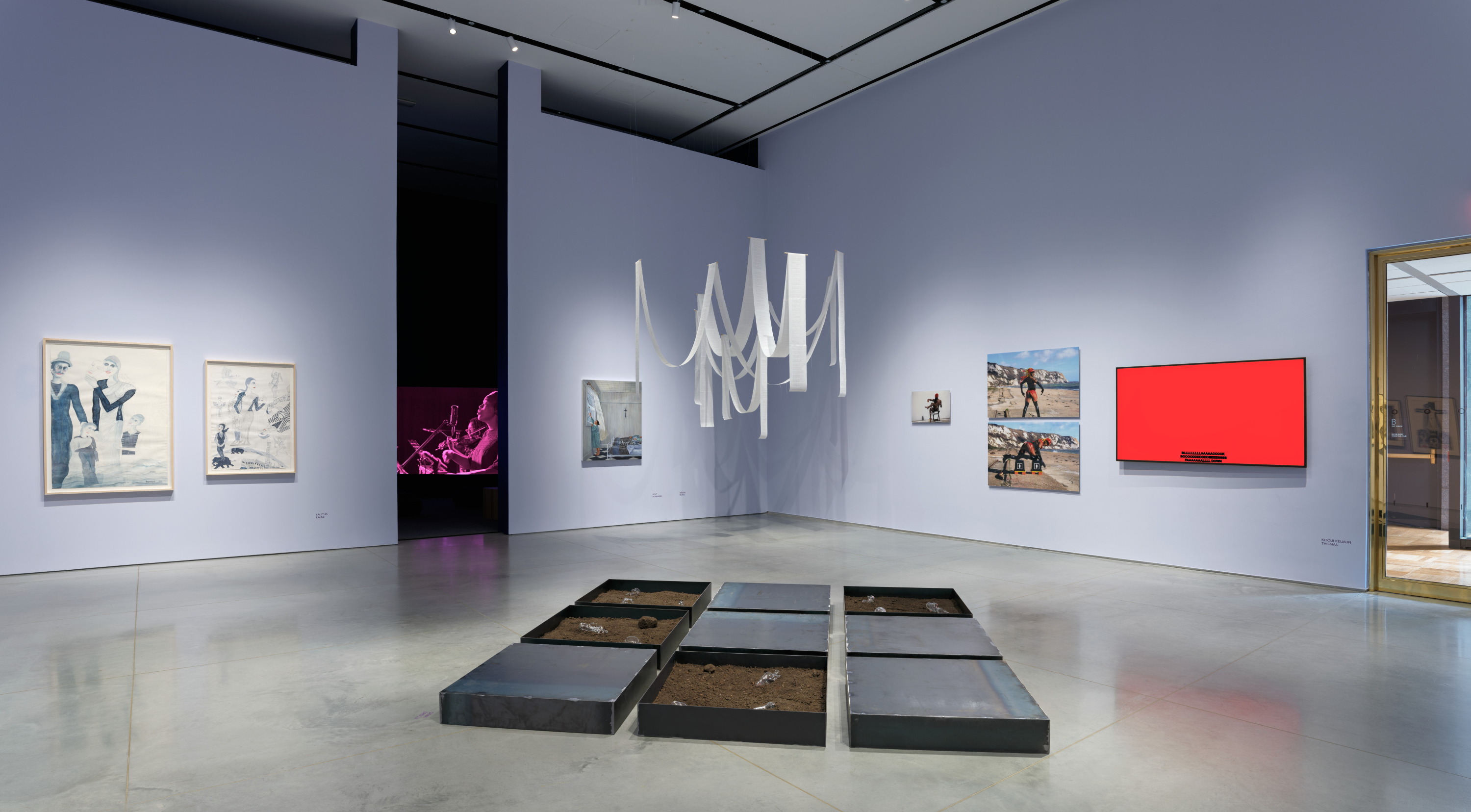 Gallery interior with a gray floor and light lavender walls with an aisle down the middle leading to a dark room with a video installation. To the right is a gold-metallic door frame. There are several artworks displayed including paintings, photographs, and a canvas mounted on the wall and a hanging white paper ribbon installation, and steel tray installation on the floor.