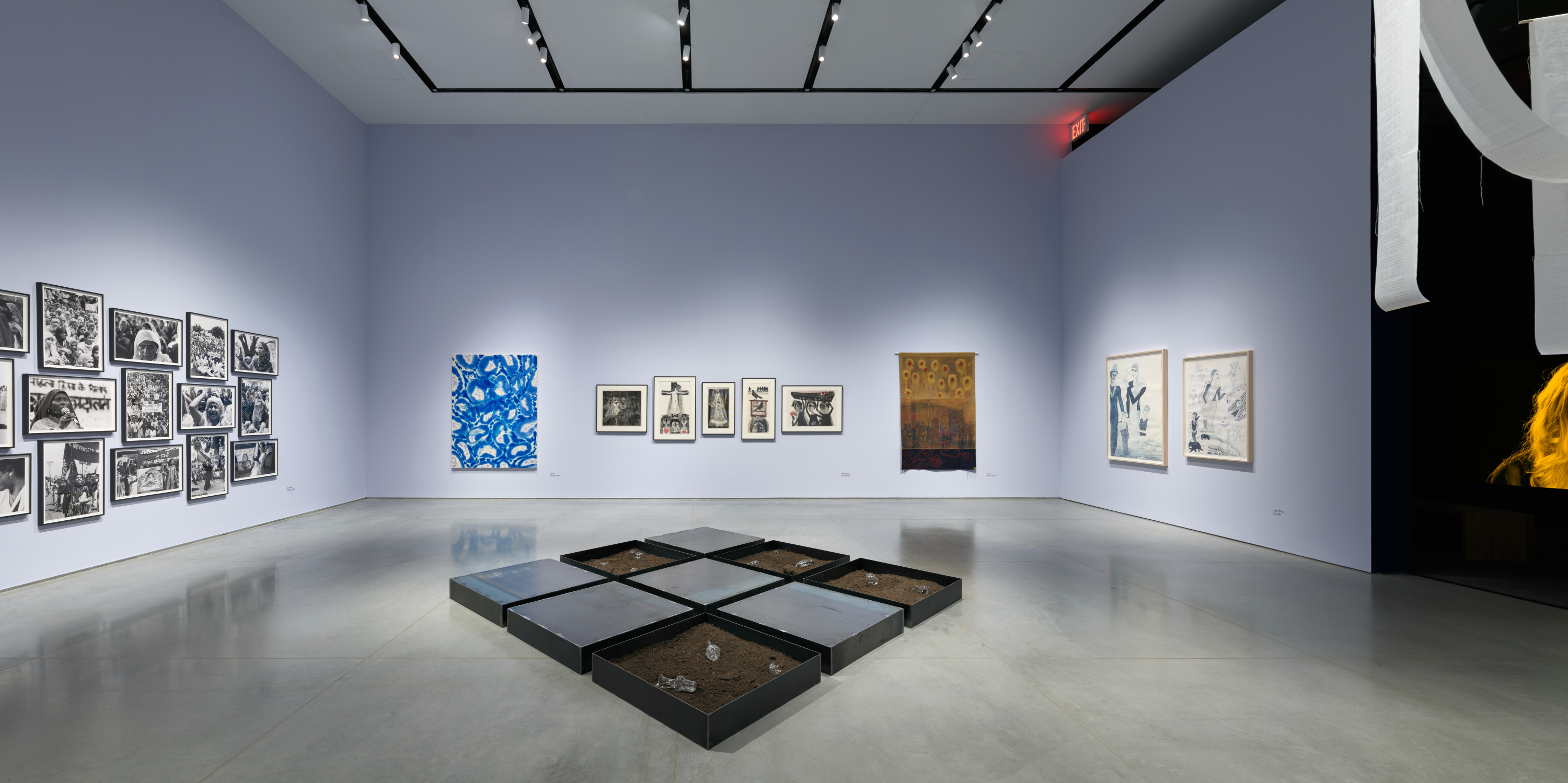 Gallery interior with light lavender walls and a gray floor. To the right is an opening to a dark room with a video installation. There are several artworks displayed including paintings, etchings, photographs, and a canvas mounted on the wall and a hanging white paper ribbon installation, and steel tray installation on the floor.