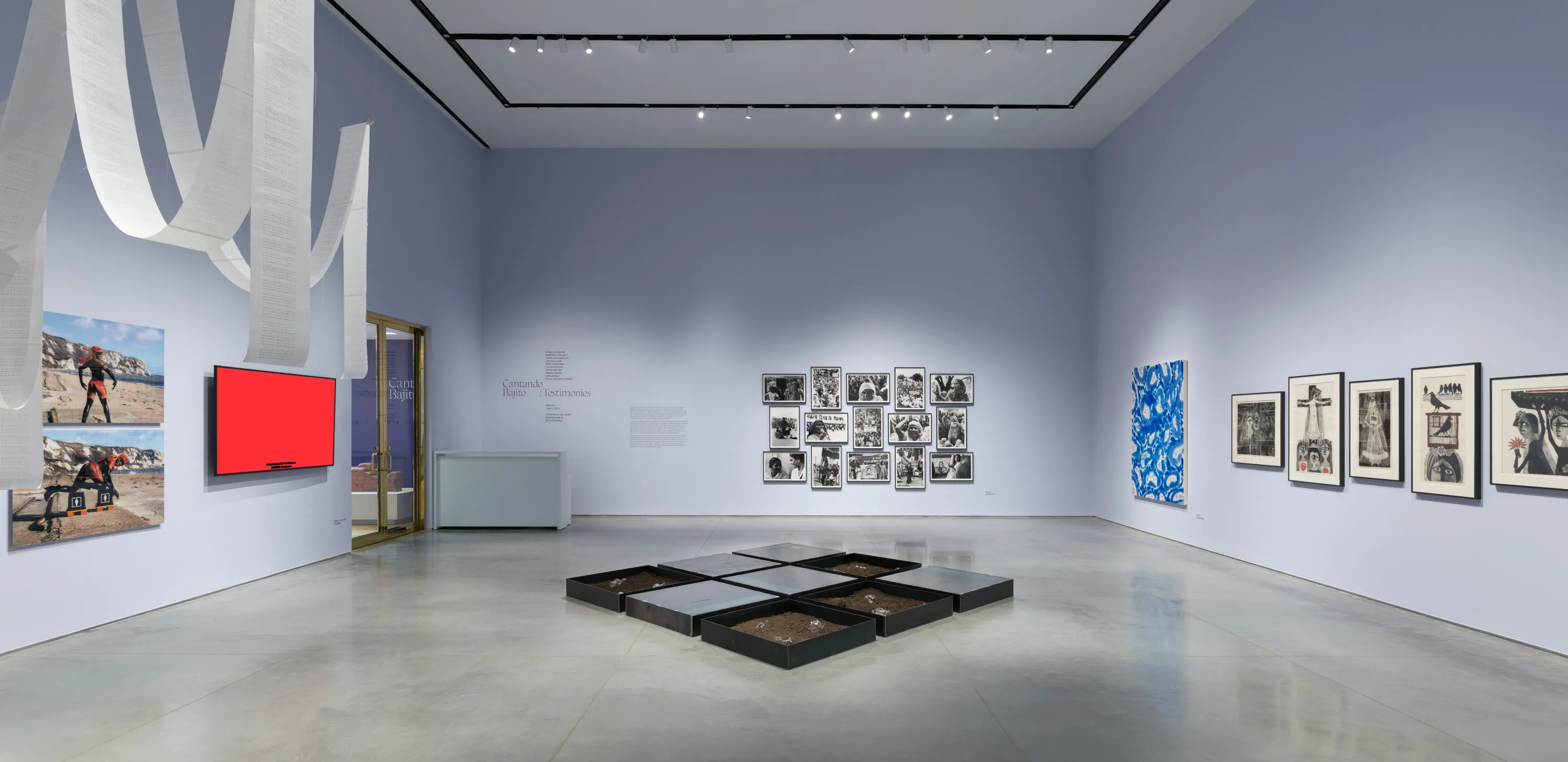 Gallery interior with light lavender walls and a gray floor. There are several artworks displayed including paintings, etchings, and photographs mounted on the wall and a hanging white paper ribbon installation, and steel tray installation on the floor.