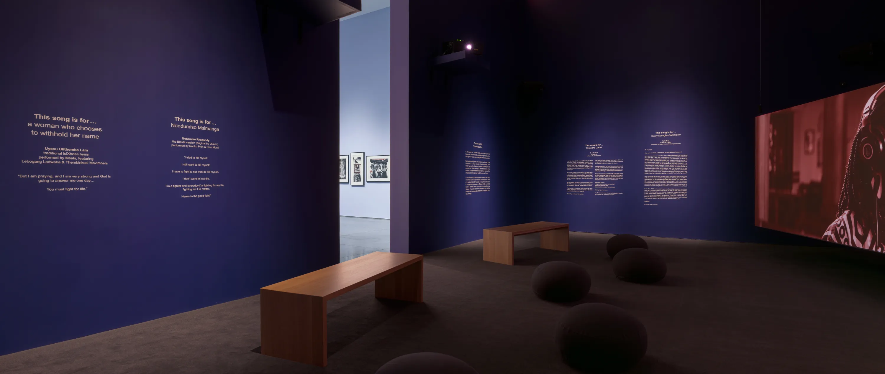 A corner of a screen in a screening room with dark purple walls and gray carpeted floor. There is a seating arrangement on the floor of two wooden benches and several cushion pods.On the walls is text relating to the artwork installation. There is a middle aisle in between the walls leading to another gallery interior with light lavender walls.