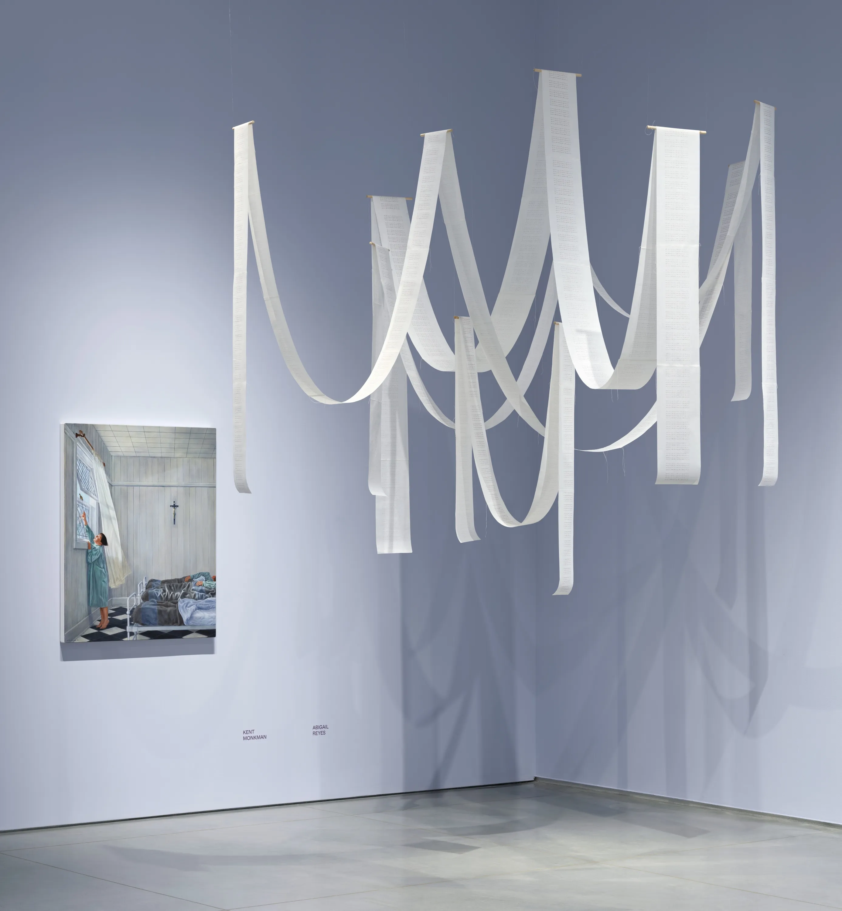 Long ribbons of white paper hanging vertically and in dipped arcs; the artwork is in front of a light lavender wall. 