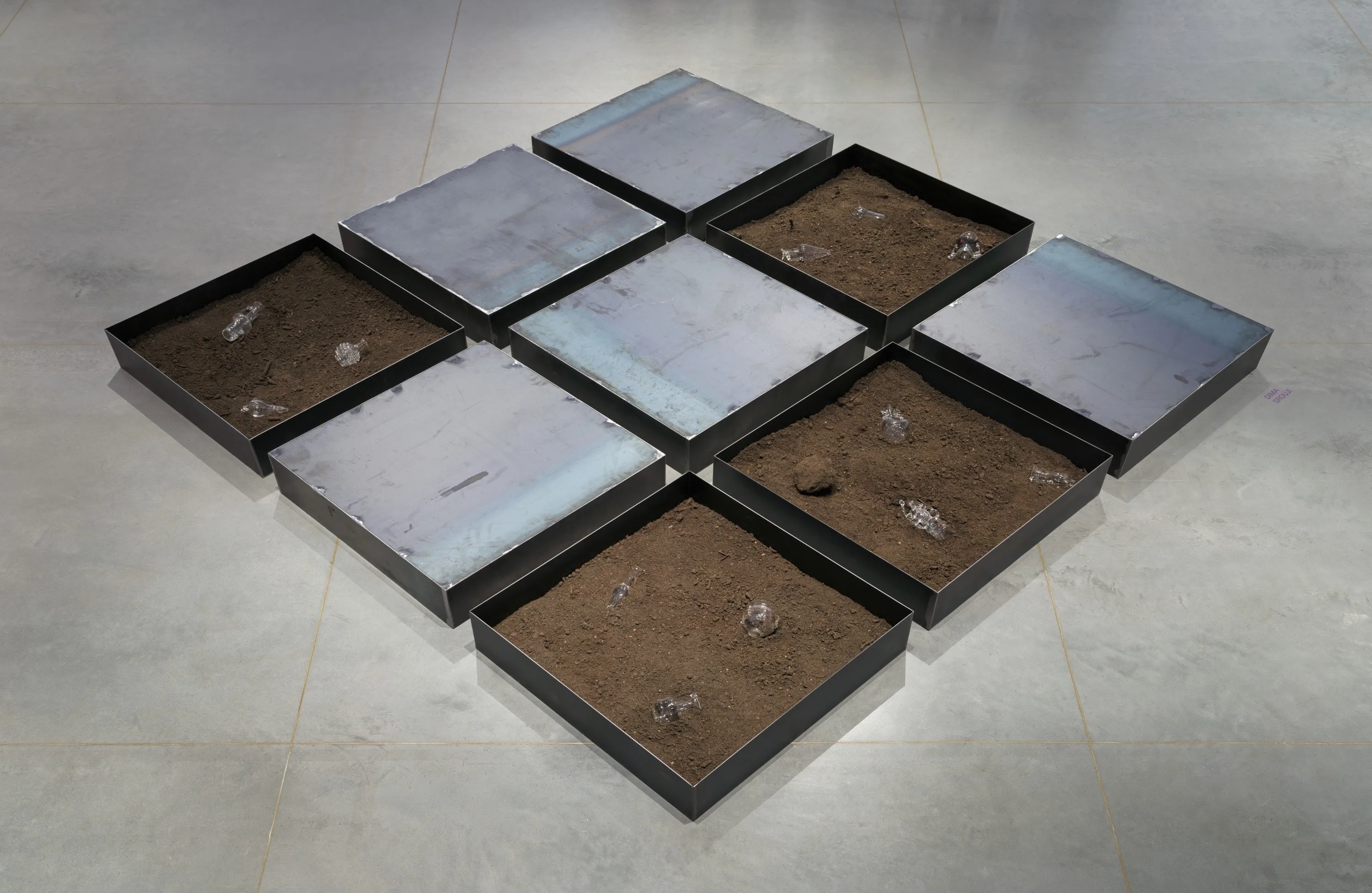 A three by three grid installation of large steel trays on the floor. Some trays are filled with dirt and glass vessels.