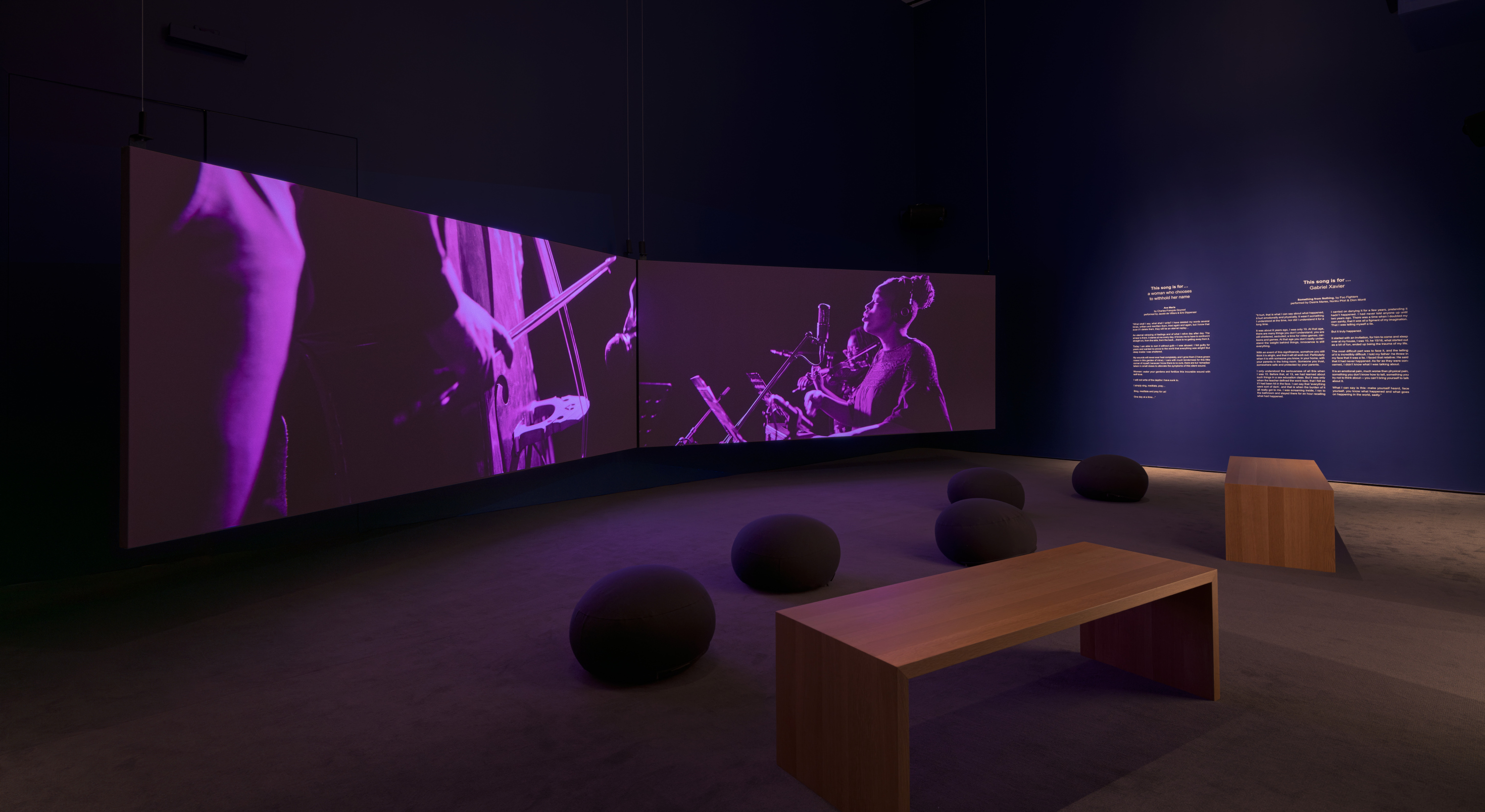 Two rectangular screens in a screening room with dark purple walls and gray carpeted floor. On the walls is text relating to the artwork installation. There is a seating arrangement on the floor of two wooden benches and several cushion pods.