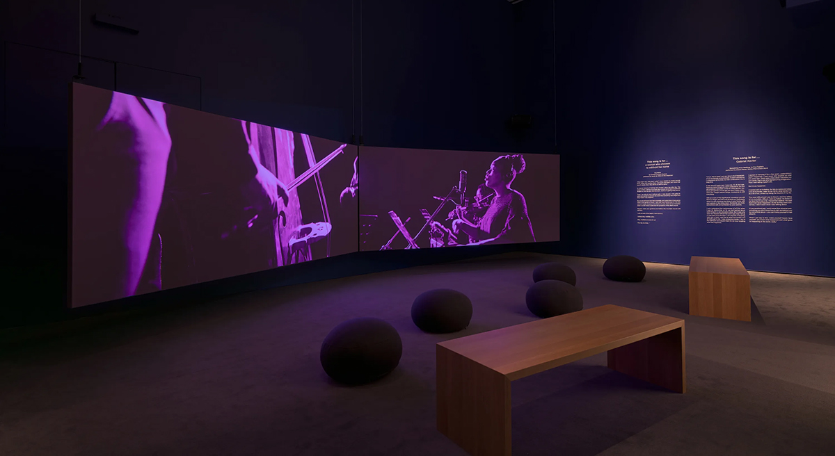 Two rectangular screens in a screening room with dark purple walls and gray carpeted floor. On the walls is text relating to the artwork installation. There is a seating arrangement on the floor of two wooden benches and several cushion pods.