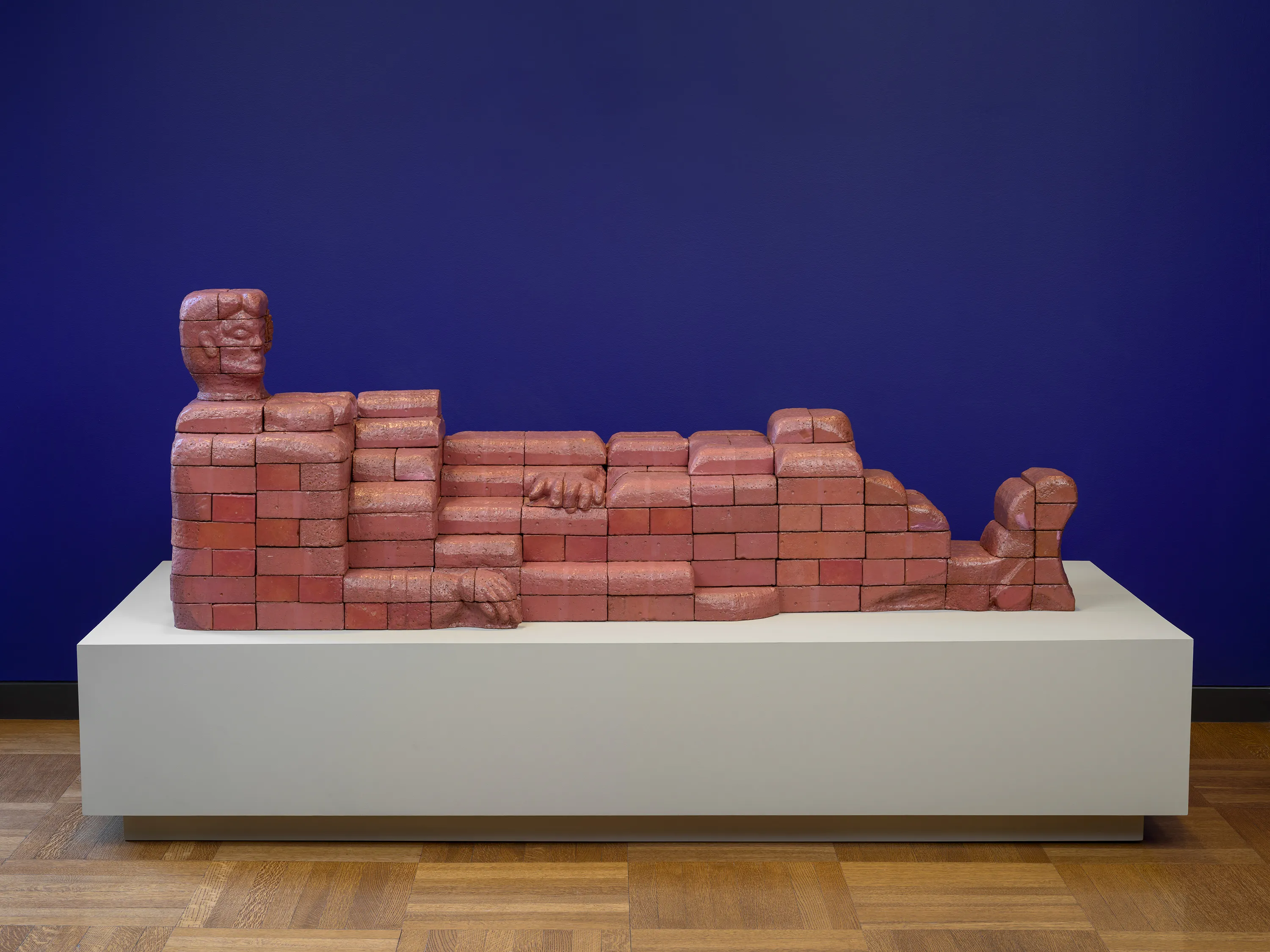 Sculpture of terracotta bricks approximating the shape of a reclining woman in front of a purple wall.