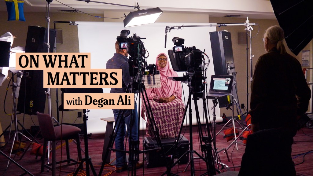 A studio setup with multiple lights, cameras, and crew members surrounding a central figure, a woman in a pink hijab and patterned dress, who is seated and being interviewed. The text overlay reads "On What Matters with Degan Ali.