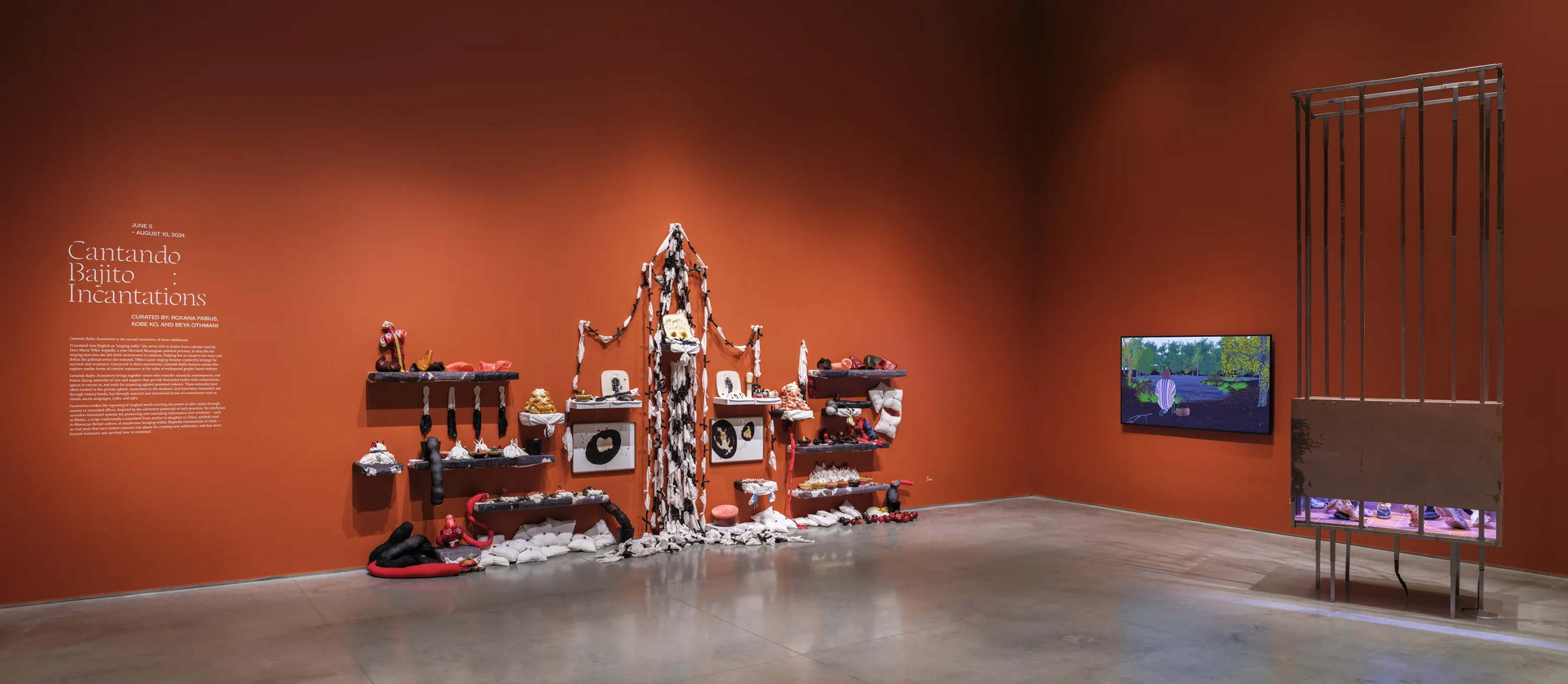 Gallery interior with terracotta-red walls and a gray floor. There are several artworks displayed - from left to right - a soft-sculpture mixed-media installation, a tv monitor with a video animation, and a ceiling-height cage sculptural installation.