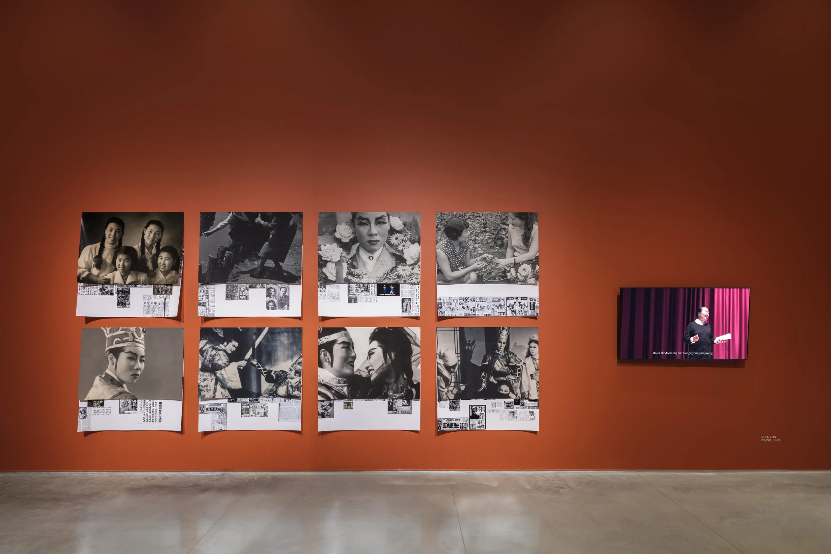 A terracotta-red wall connecting to a gray floor. On the left side of the wall is a 2-row and 4-column photo collage featuring historical photographs of East Asian stage performers and Korean newspaper cuttings. To the right is a tv monitor featuring a video of an East Asian woman on a stage giving directing notes in Korean.