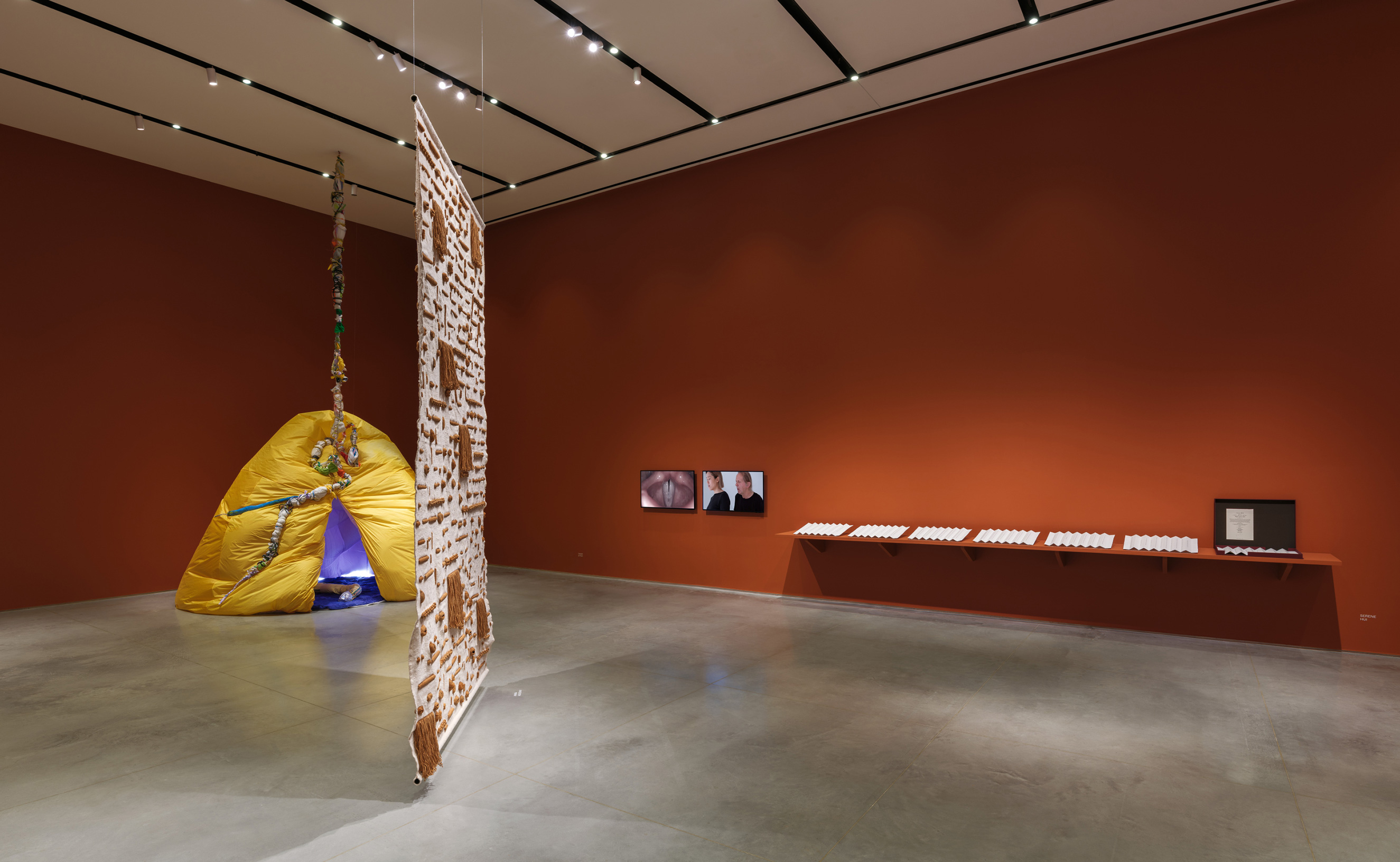 Gallery interior with terracotta-red walls and a gray floor. Suspended from the ceiling in the middle of the gallery floor is a sand-colored tapestry with brown geometric details. To the left is an inflatable yellow hut, split in the middle with an emanating purple light. And on the right wall is two video monitors and a shelf with a row of embossed papers.